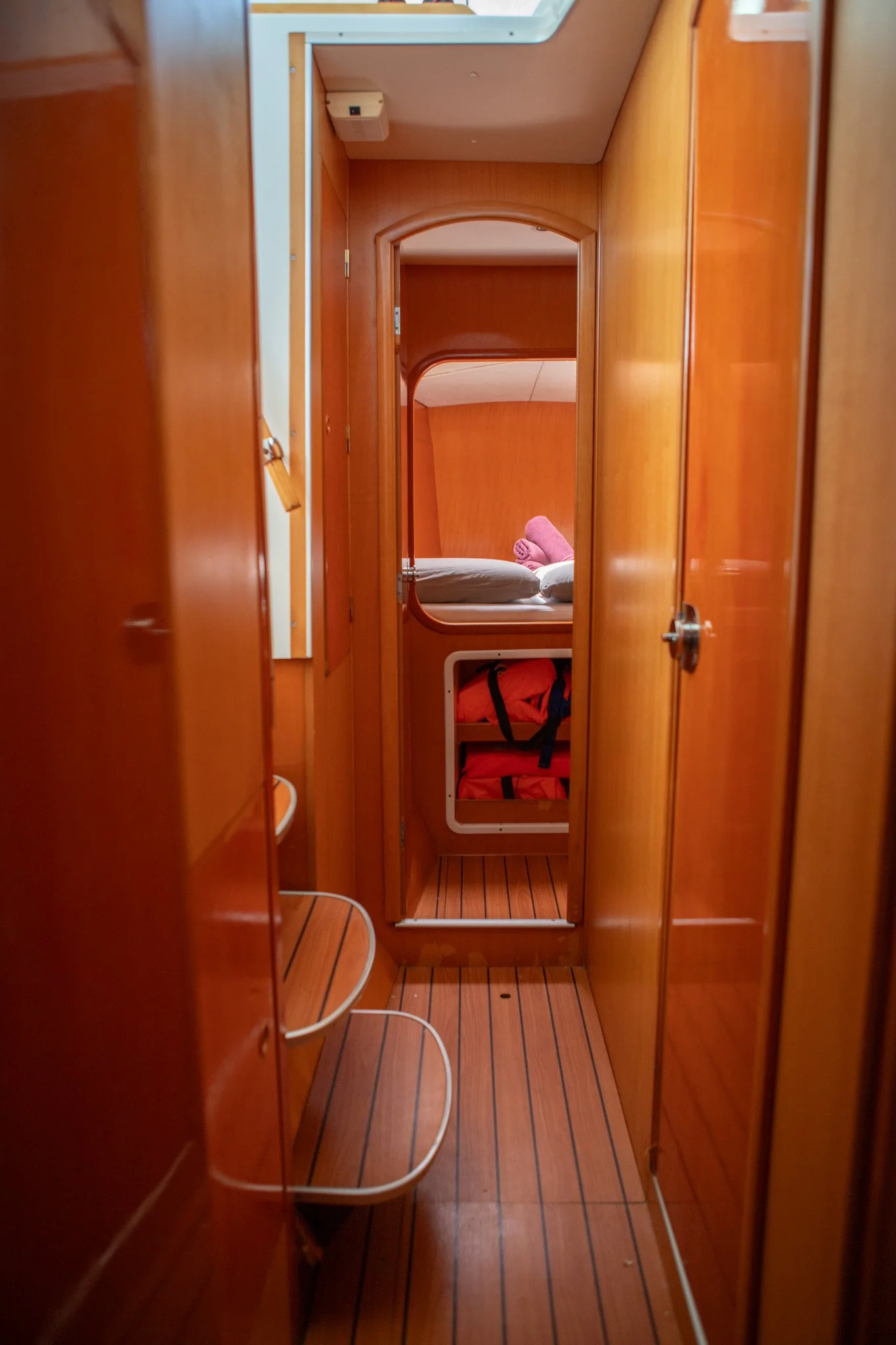View of a narrow wooden corridor inside a vessel or boat with a bed at the end, held with pink towels and pillows, visible through a mirror. The corridor has wooden floors and walls, with white trim and a small storage compartment on the left.