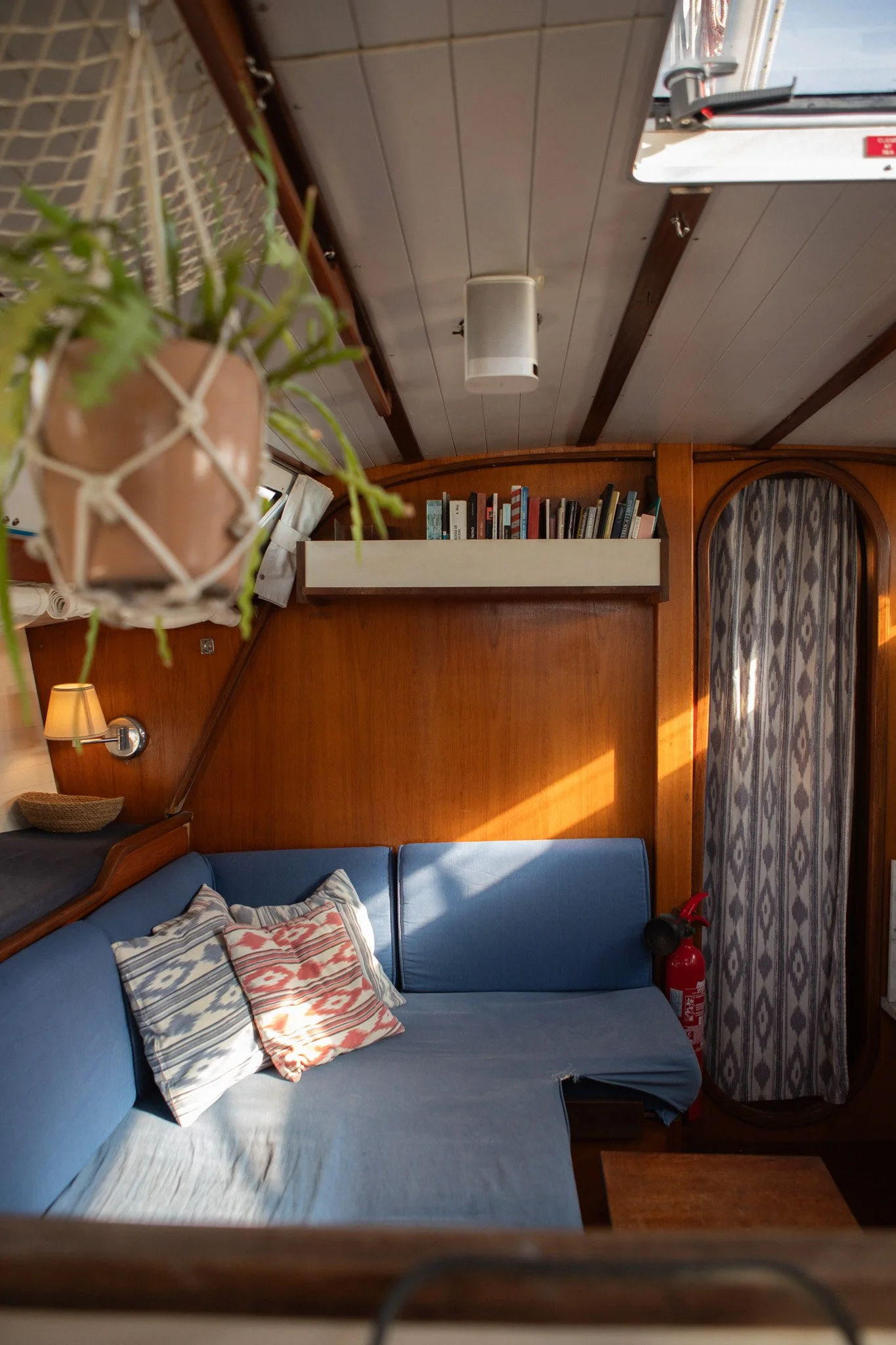 Interior of a boat cabin with a blue couch, patterned pillow, wooden wall with bookshelves, a small window with curtains, a fire extinguisher, and a potted plant hanging from the ceiling.