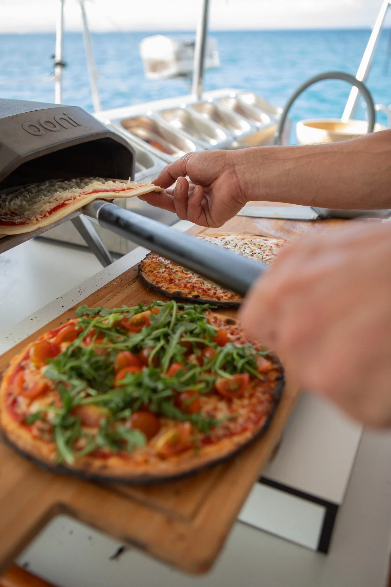 Person placing a pizza with arugula and vegetables into a pizza oven on a boat, with the ocean in the background.