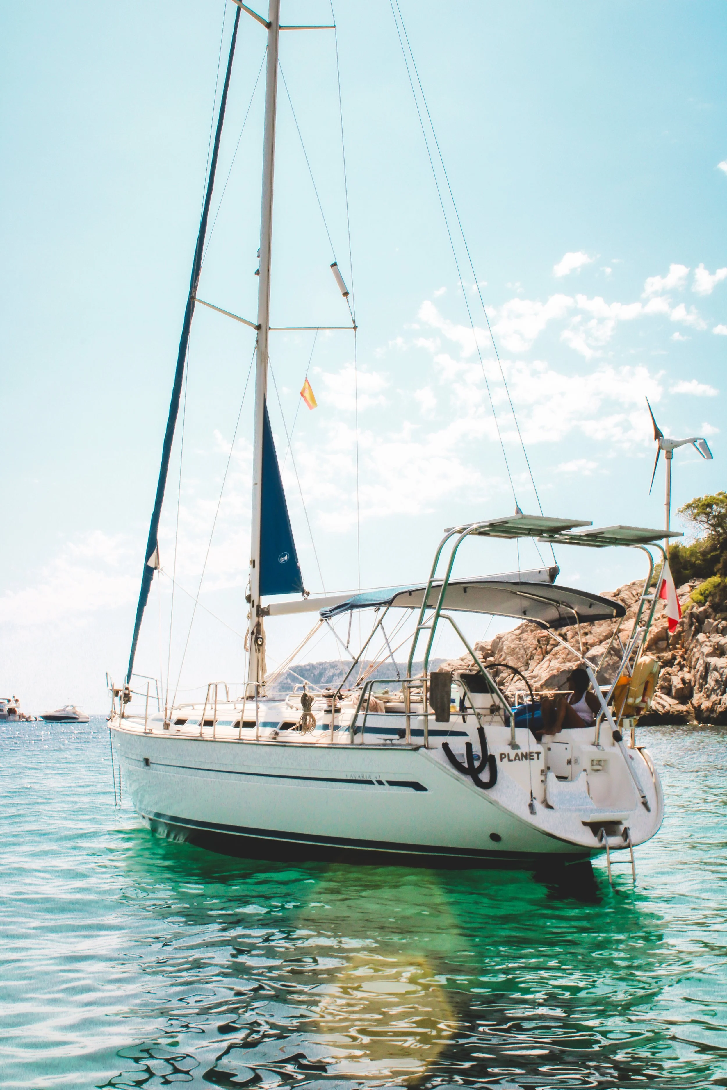 A white sailboat named 'PLANET' floating in turquoise water with multiple other boats in the distance. There are rocky hills and a wind turbine in the background, under a clear blue sky with some scattered clouds.