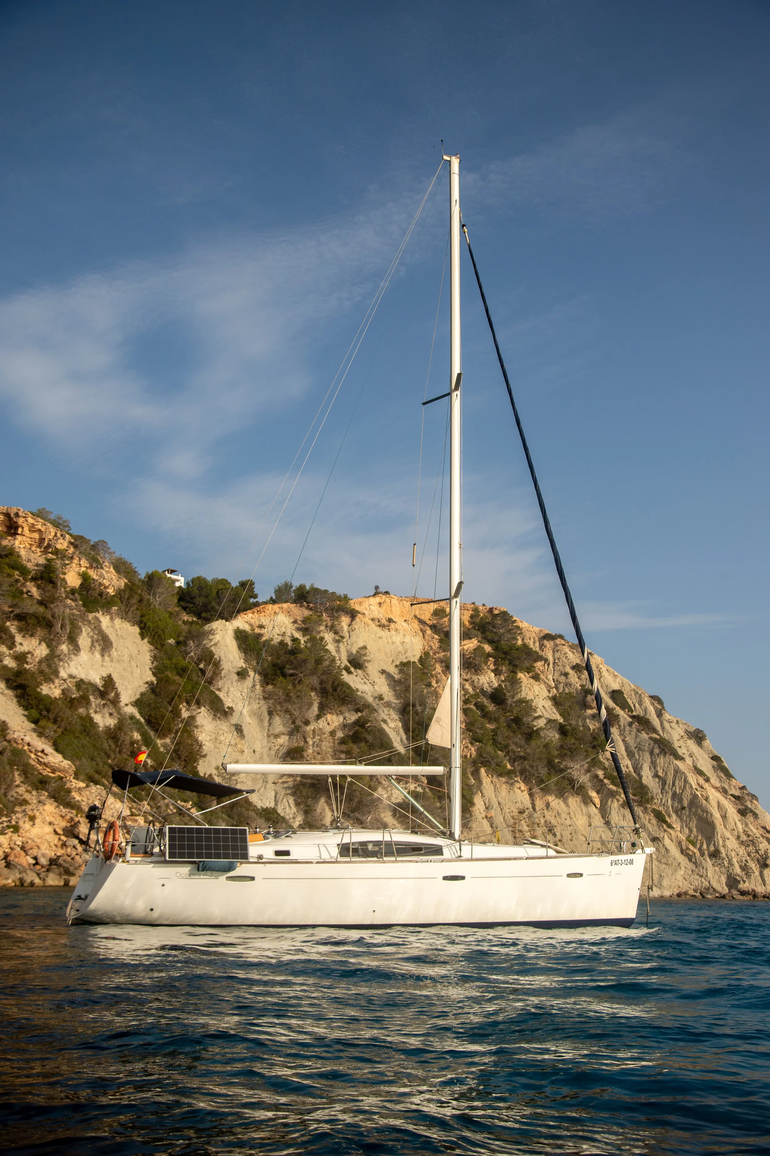 White sailboat with solar panel on deck floating on the water near rocky cliffs and hills with trees, under a clear blue sky.