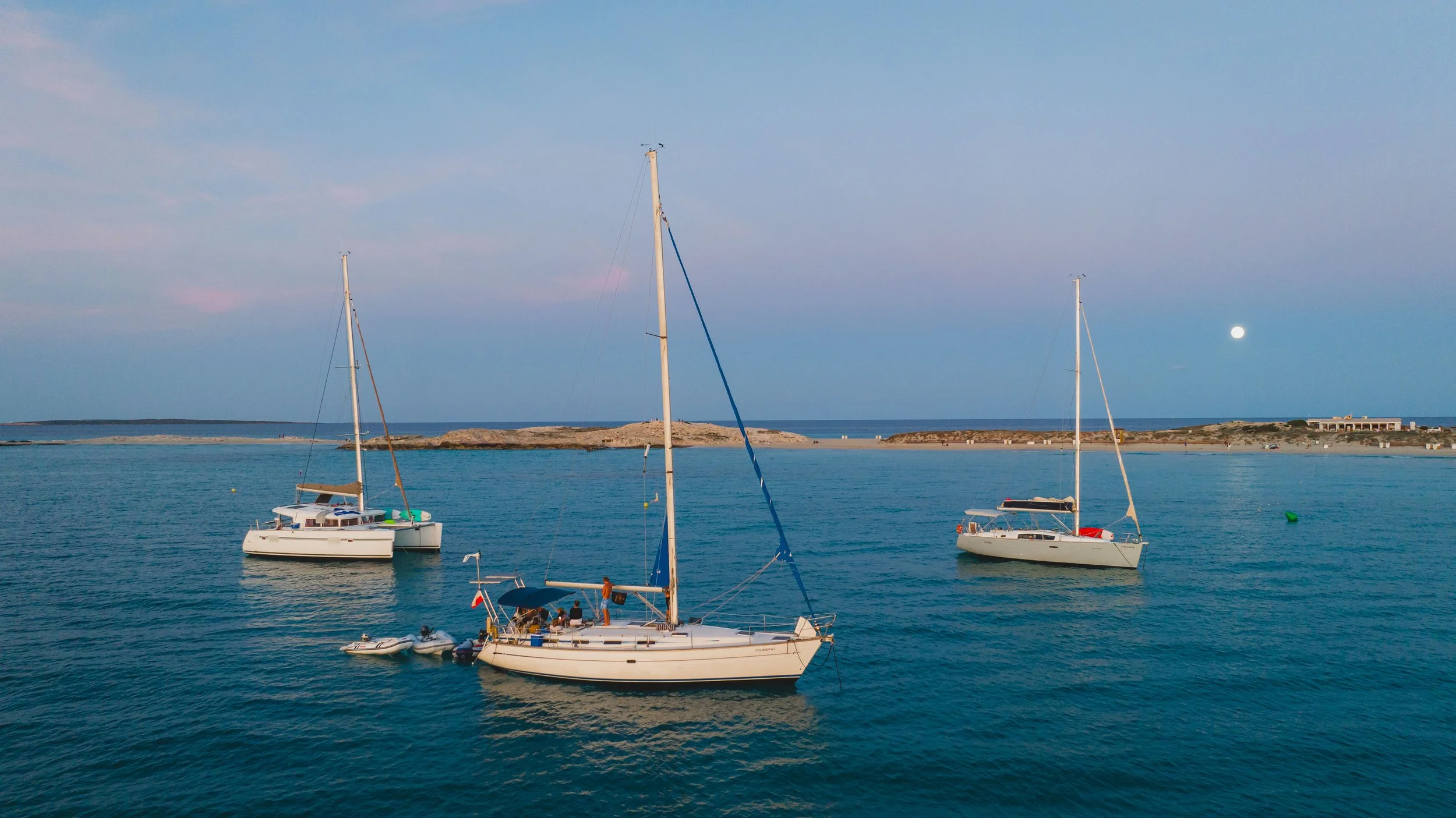 Sailboats anchored in calm blue water near a rocky shoreline with a building, under a clear sky at dusk or dawn.