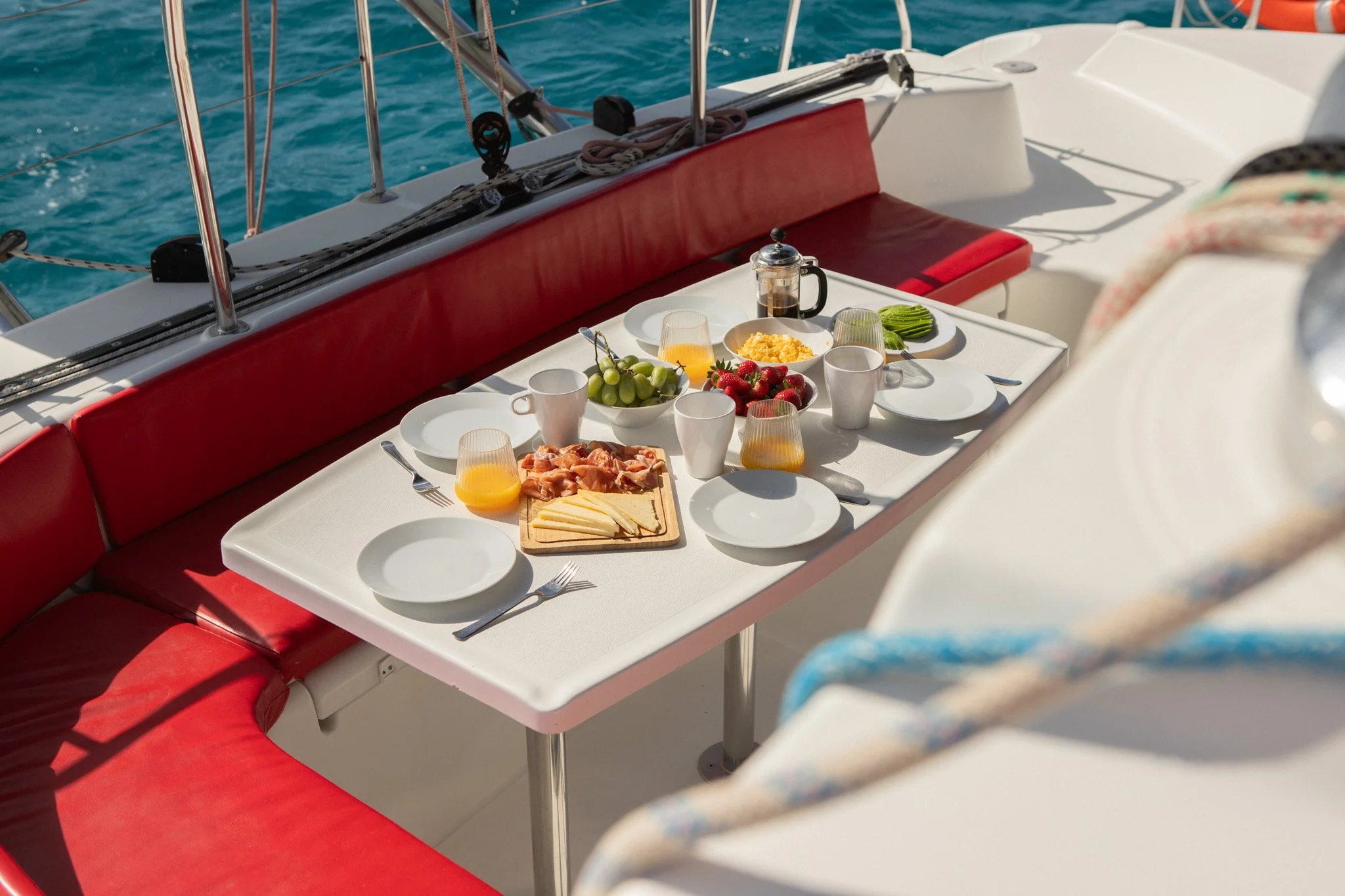 Breakfast setup on a yacht with a white table, red cushioned seating, and food including strawberries, grapes, sliced cheese, ham, orange juice, coffee, and green napkins, with the ocean in the background.