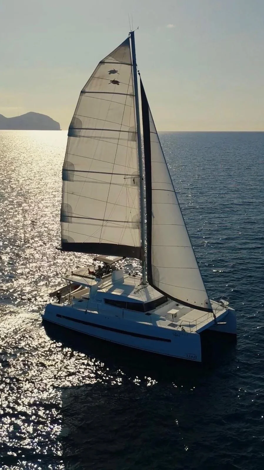 A sailboat with large sails on the ocean during sunset, with land in the distance.
