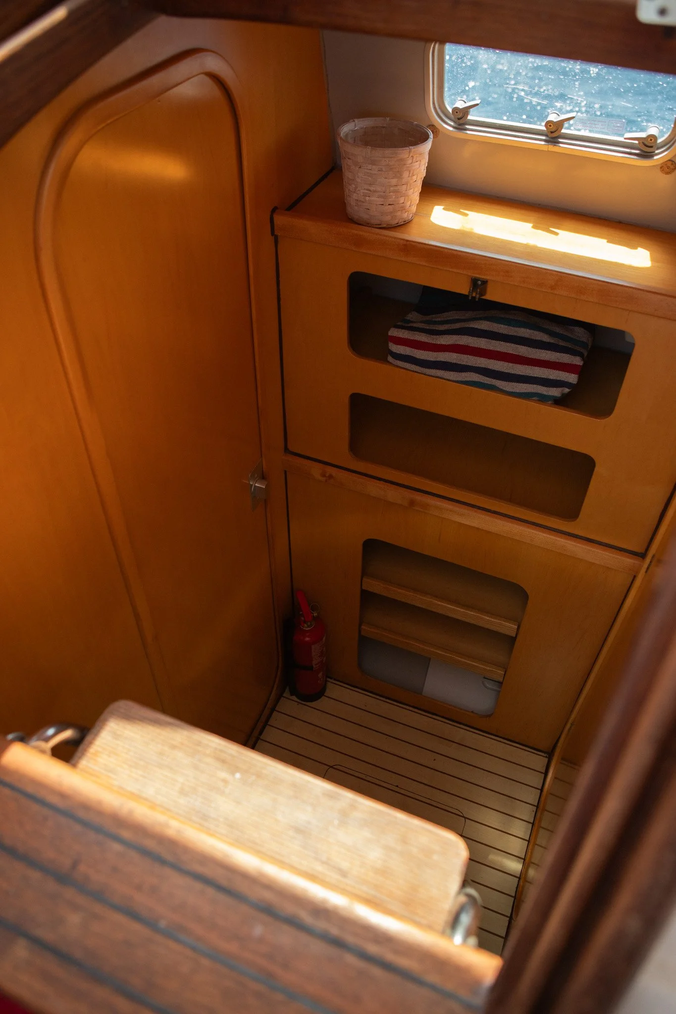 Interior of a wooden boat cabin showing a small window with water outside, a woven basket on a shelf, a towel in a cubby, and a fire extinguisher at the bottom corner.