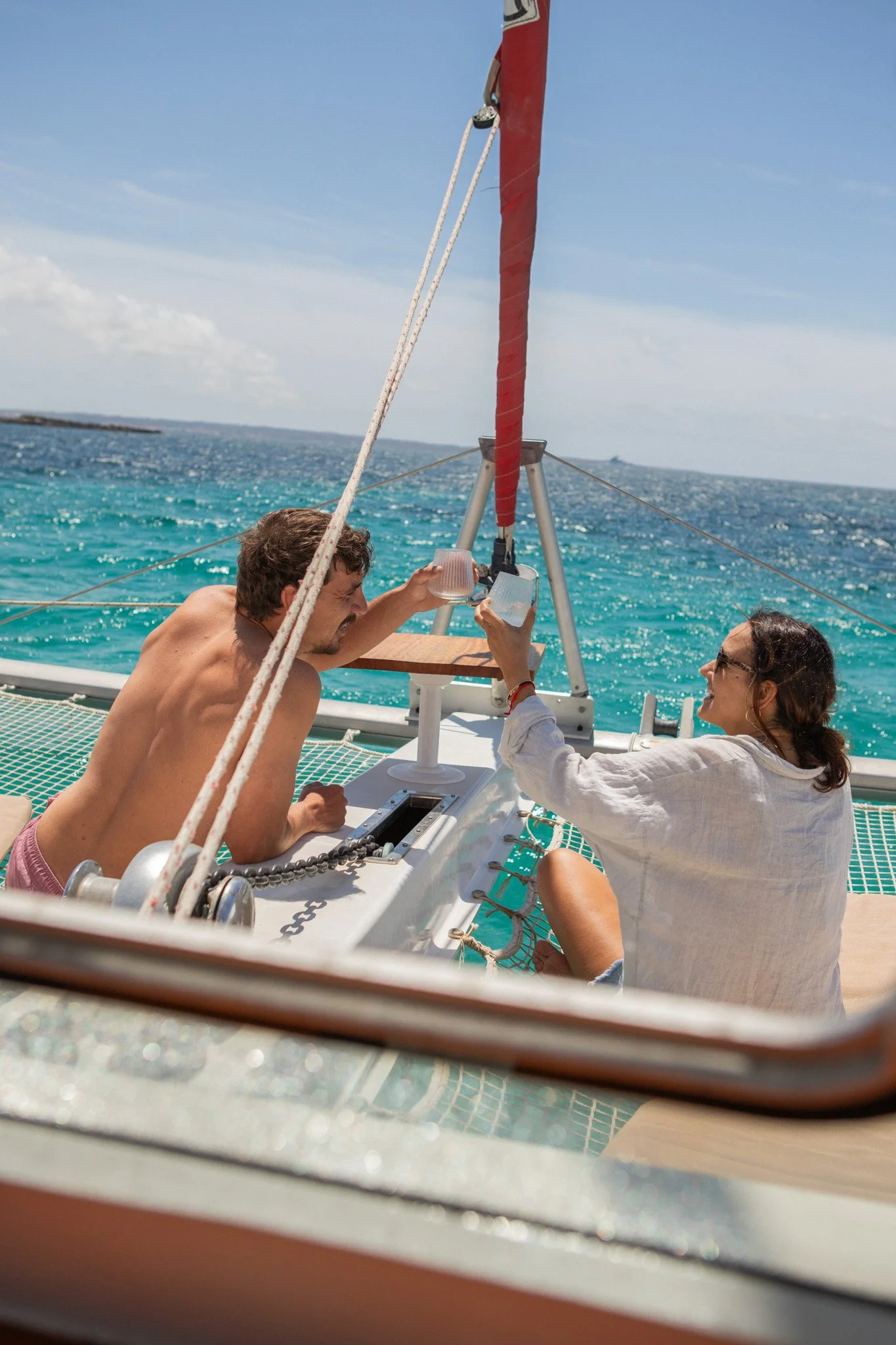 Two people sitting on a boat with a sail, enjoying the ocean, taking a photo, under a partly cloudy sky.
