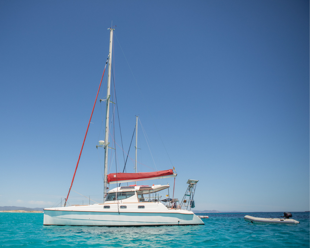 A sailboat with a white hull and red sail cover floating on turquoise water near a small inflatable boat.