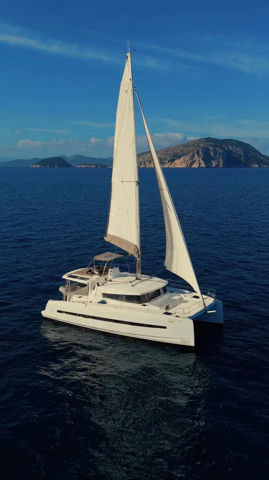 A white sailing yacht with tall sails on calm deep blue water, with mountainous islands in the background under a clear blue sky.