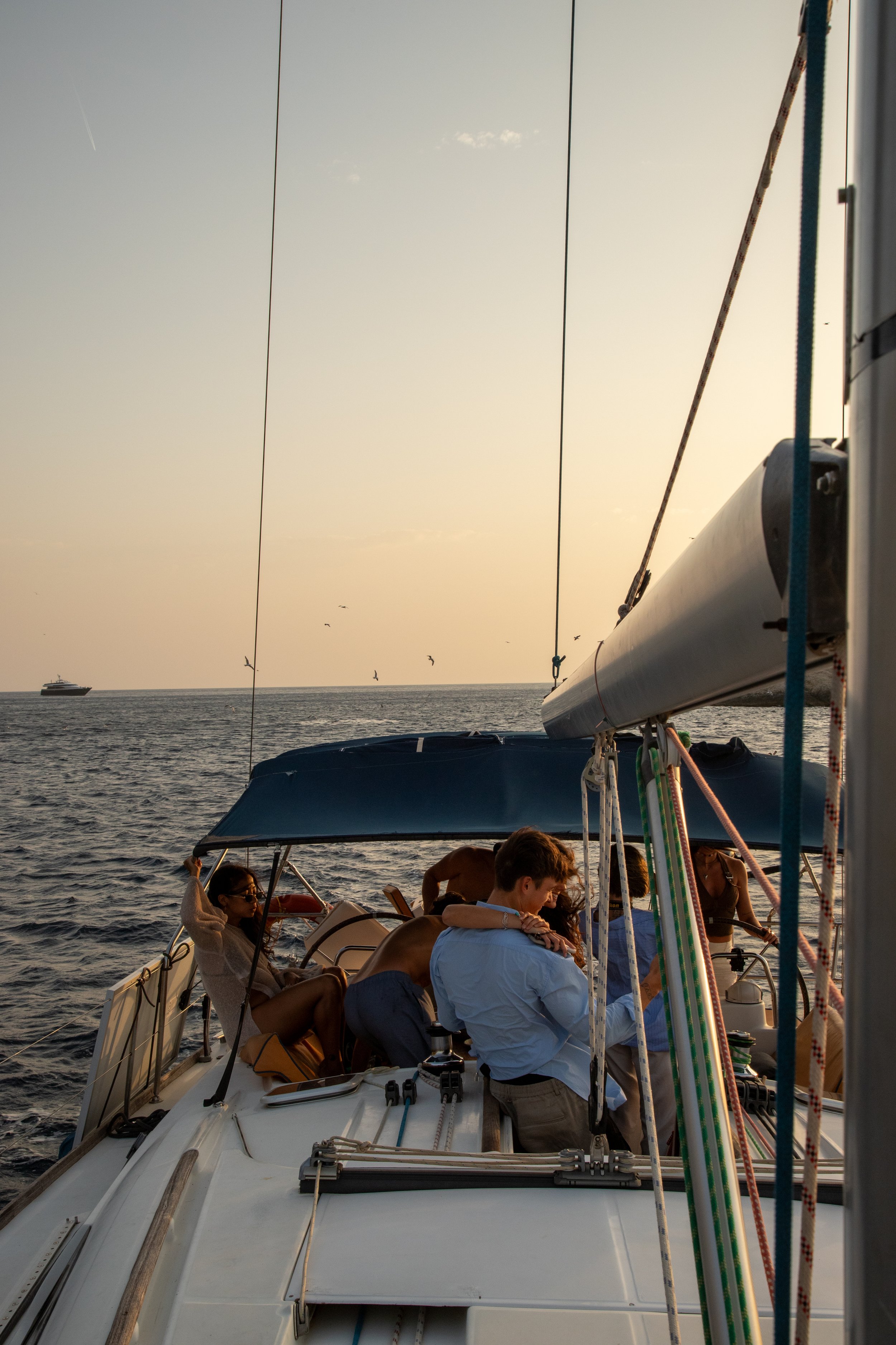 People relaxing and socializing on a sailboat during sunset, with a view of the open water and distant ship, some individuals wearing swimsuits and sunglasses.