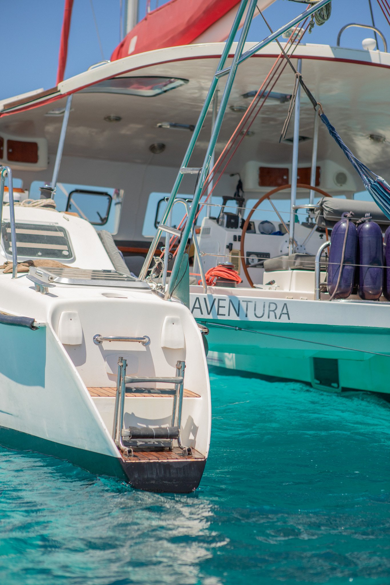Close-up view of sailboats anchored in turquoise water, with one boat named 'Aventura' visible. The boats have red and white sails, equipment, and safety gear on deck, under a clear blue sky.