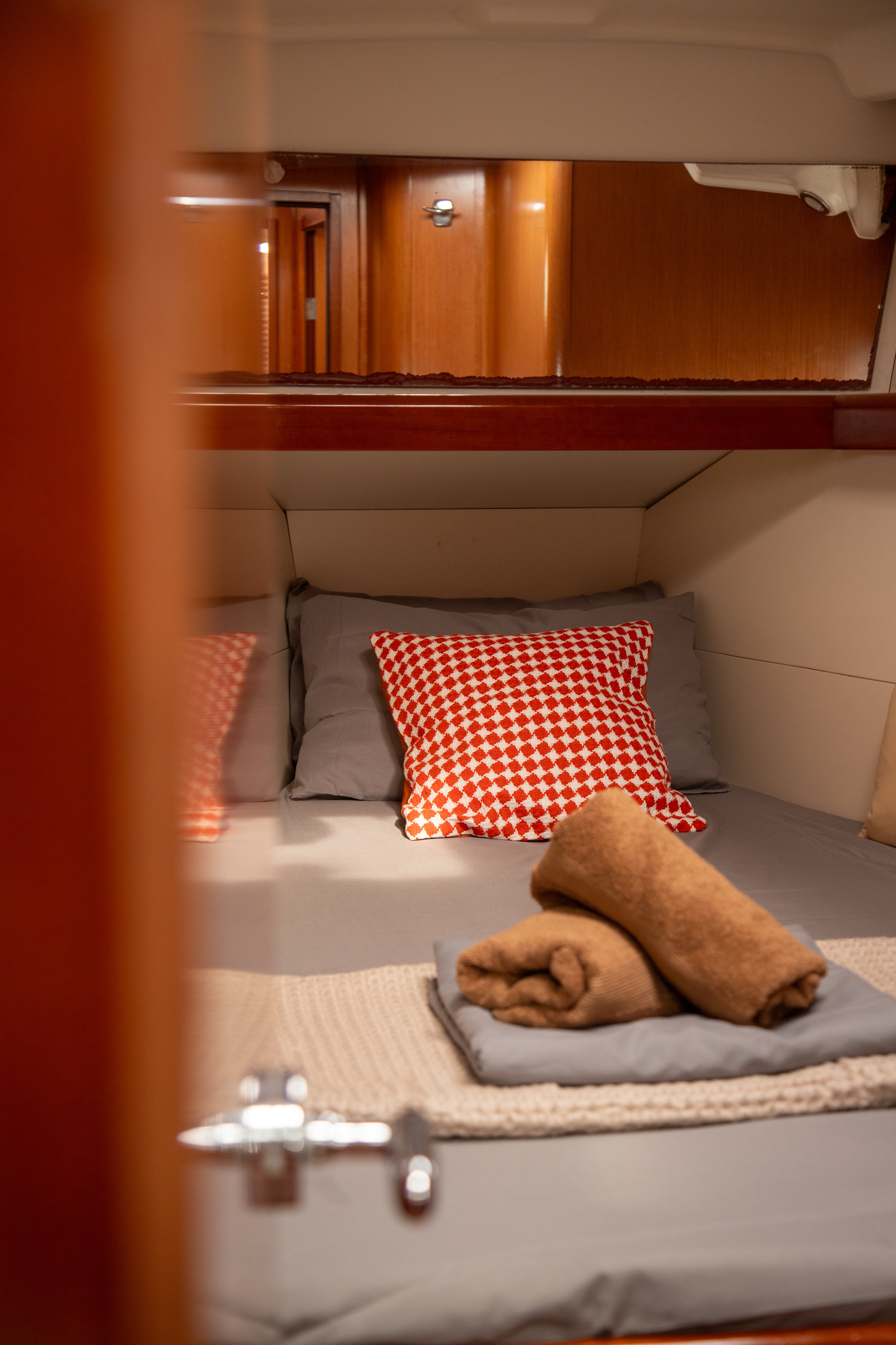 View of a cozy bed in a small cabin or RV, with gray bedsheets, a red and white checkered pillow, and a rolled-up beige towel on top of the bed, seen through a partially open door.