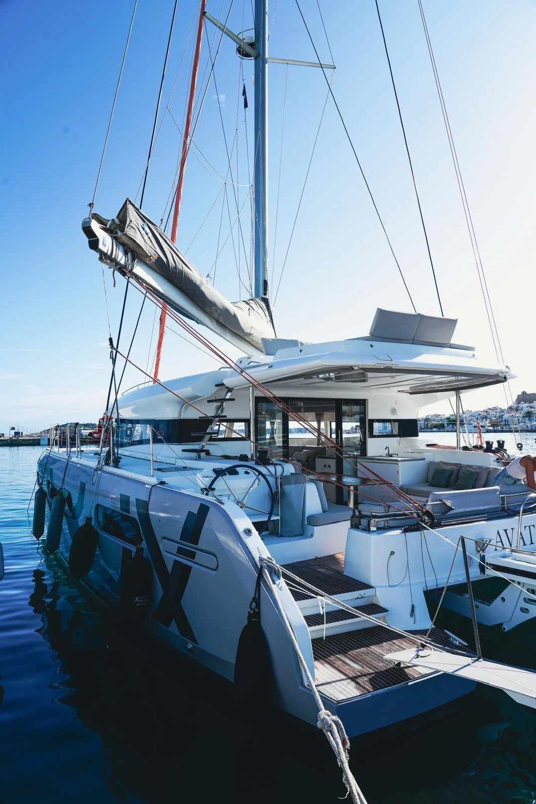 View of a modern white sailboat docked at a marina on a clear day with blue skies, showing the deck, mast, and water reflections.