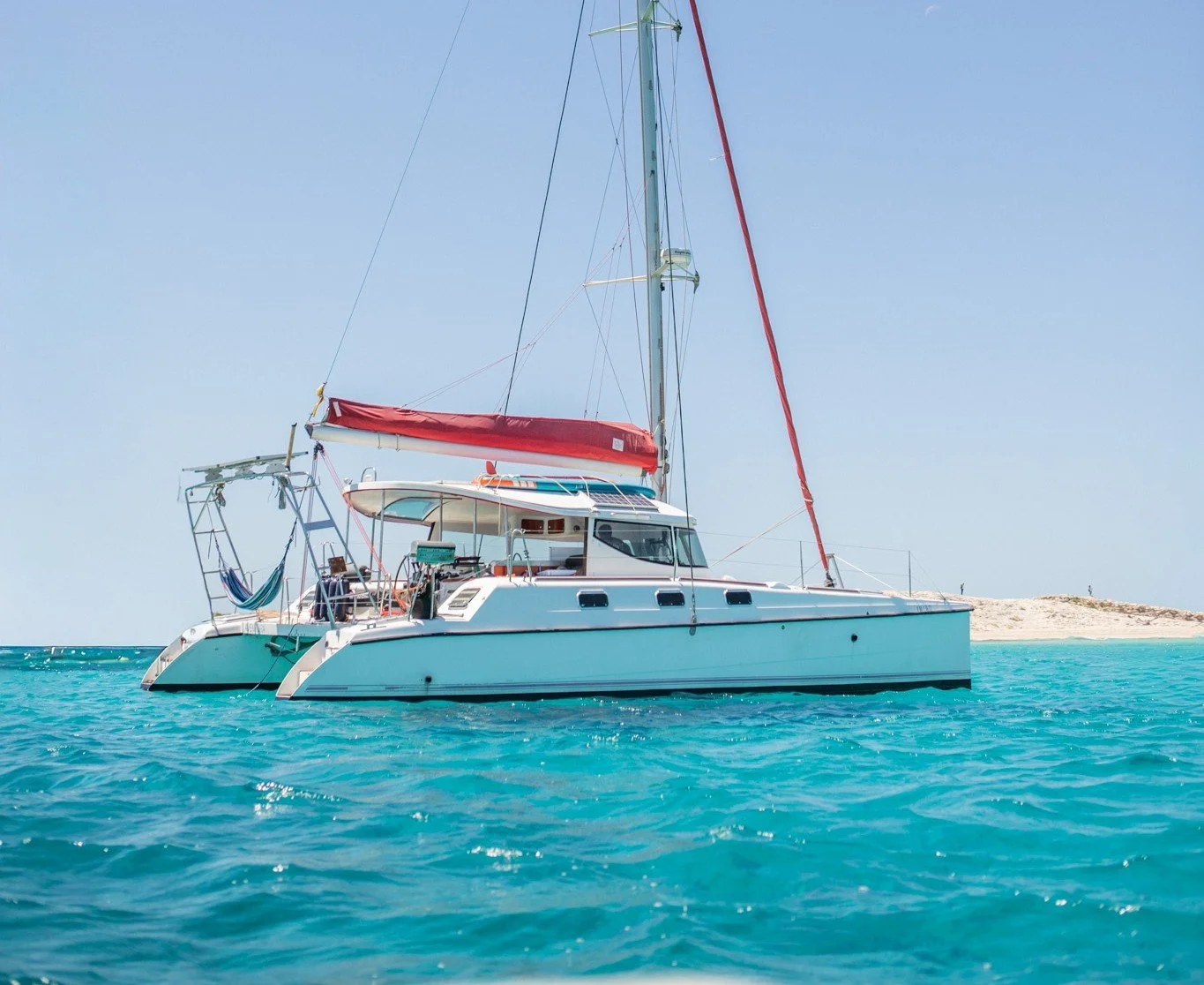 A white sailing catamaran yacht with a red sail cover floating on turquoise water near a small island with sandy shores and low vegetation, under a clear blue sky.
