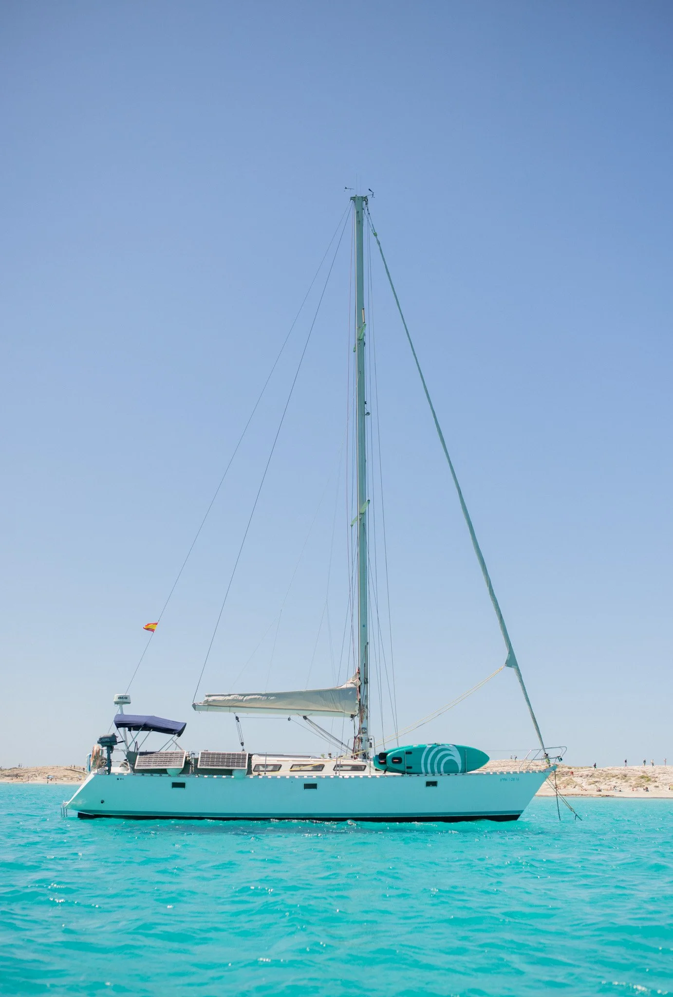 A white sailboat with various equipment and solar panels sails on clear turquoise water near a sandy shoreline, under a clear blue sky.