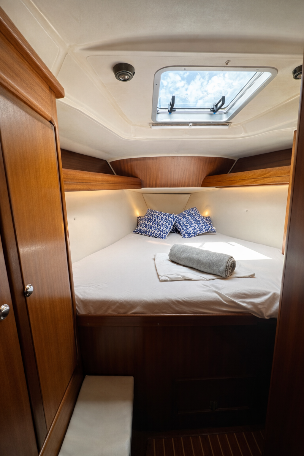 A cozy boat cabin bedroom with a bed, blue patterned pillows, a rolled gray towel, and a view of the sky through a ceiling hatch.