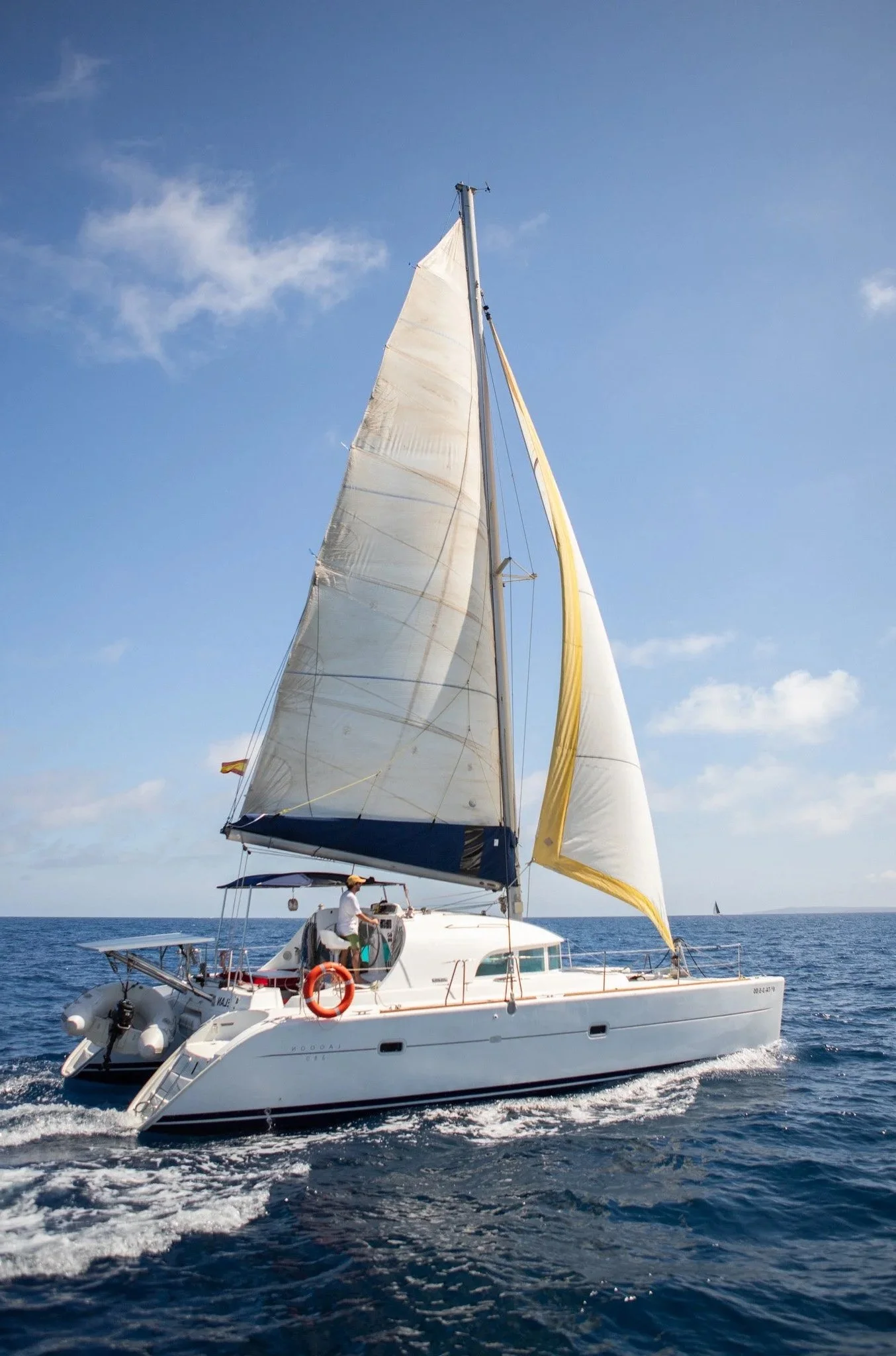 A large white sailing yacht with blue accents moving through open water under a blue sky with some clouds. The yacht has its sails up, including a yellow sail. A person in a white shirt and hat is on the deck.