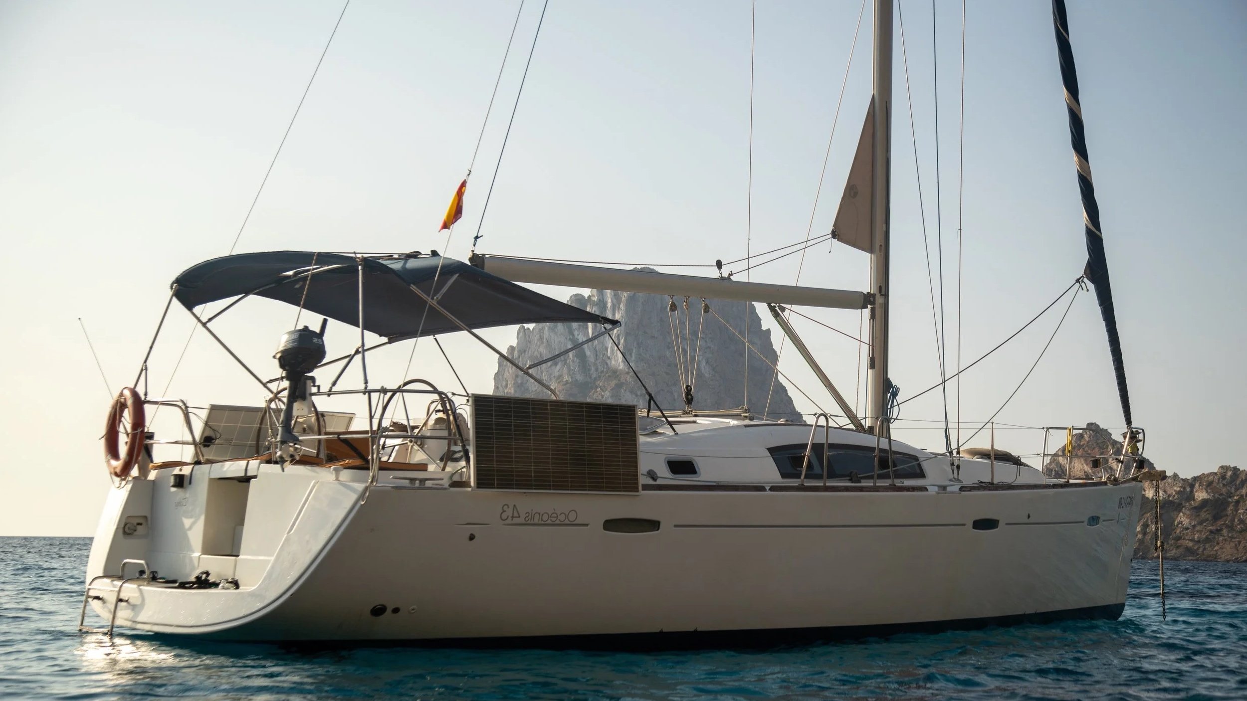 A sailing yacht on the water with a rocky island in the background, under a clear sky.