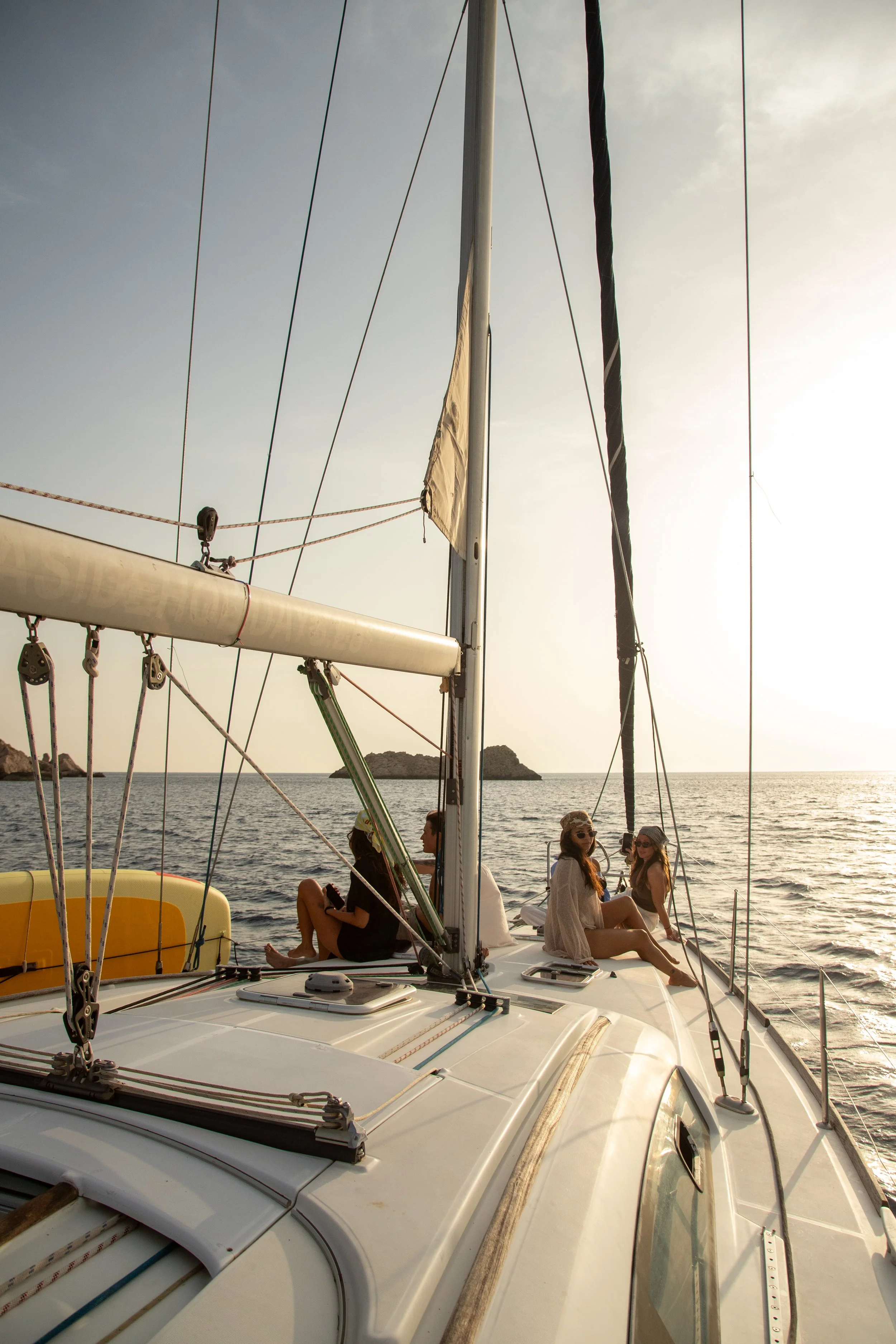 Three women sitting and chatting on a sailboat, with a distant view of the sea and small islands in the background during sunset.