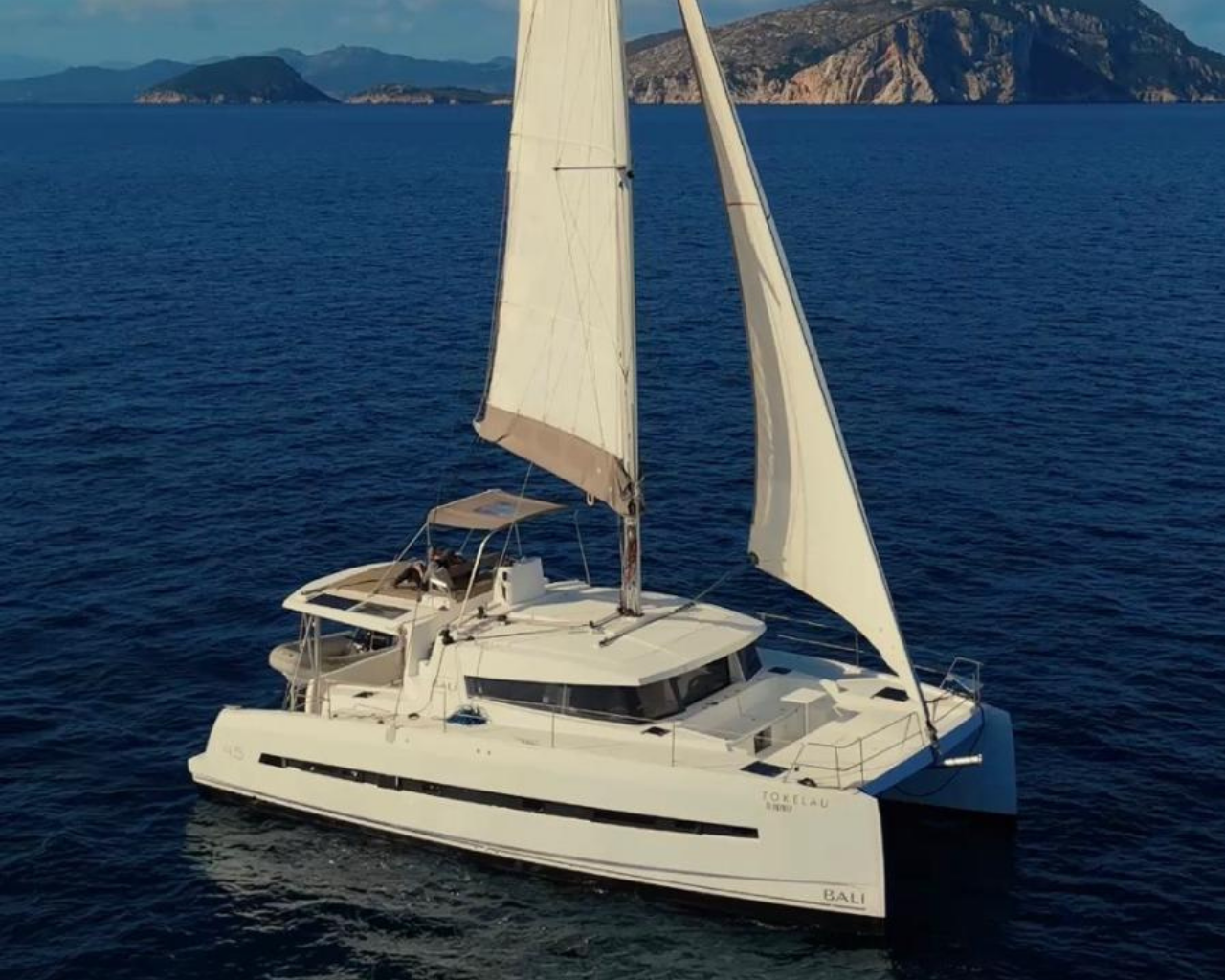 A white catamaran sailing yacht named Bali on calm blue water with an island and mountains in the background.