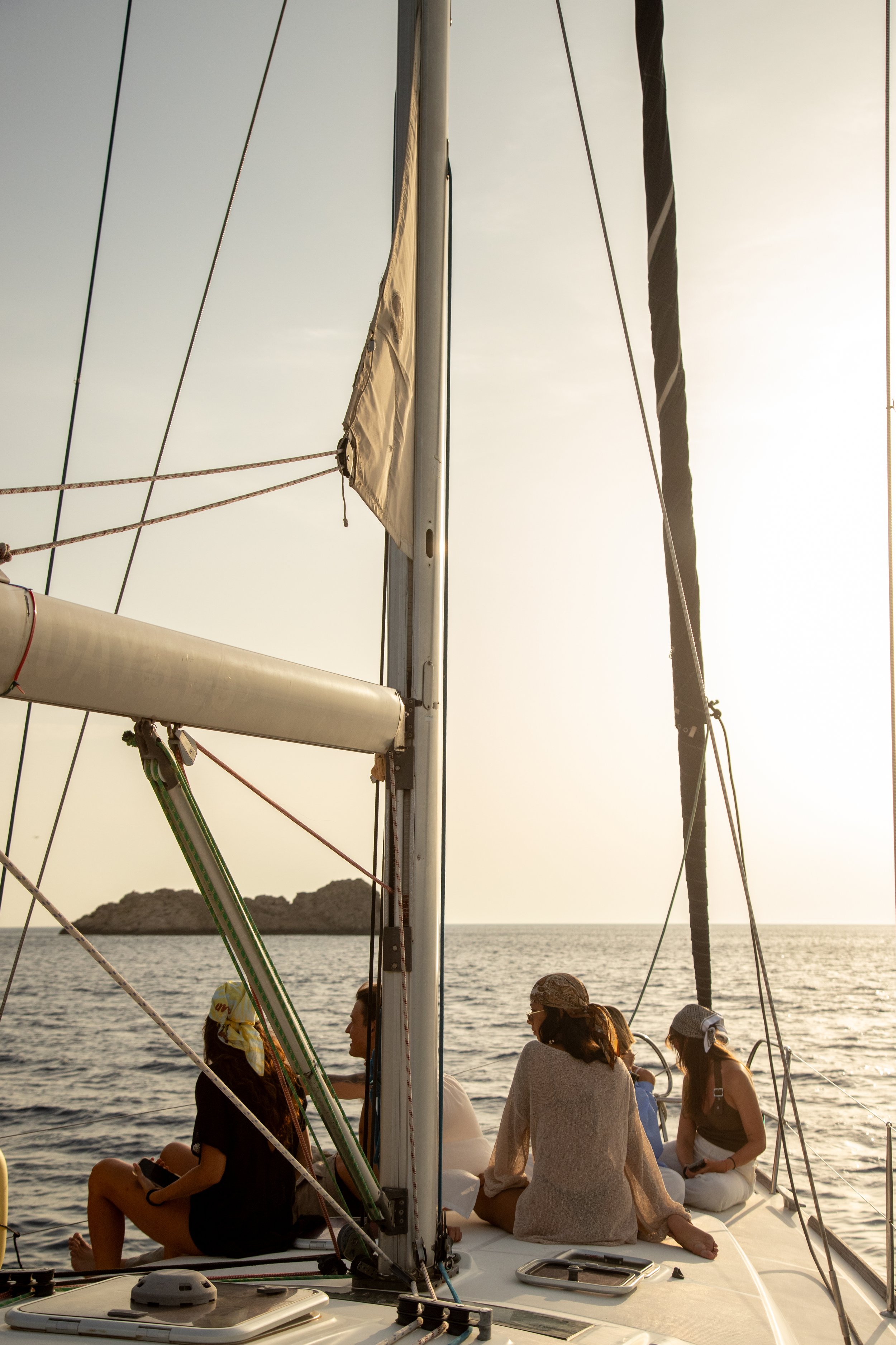 People sitting and talking on a sailboat during sunset, with a small island visible in the background and a clear sky.