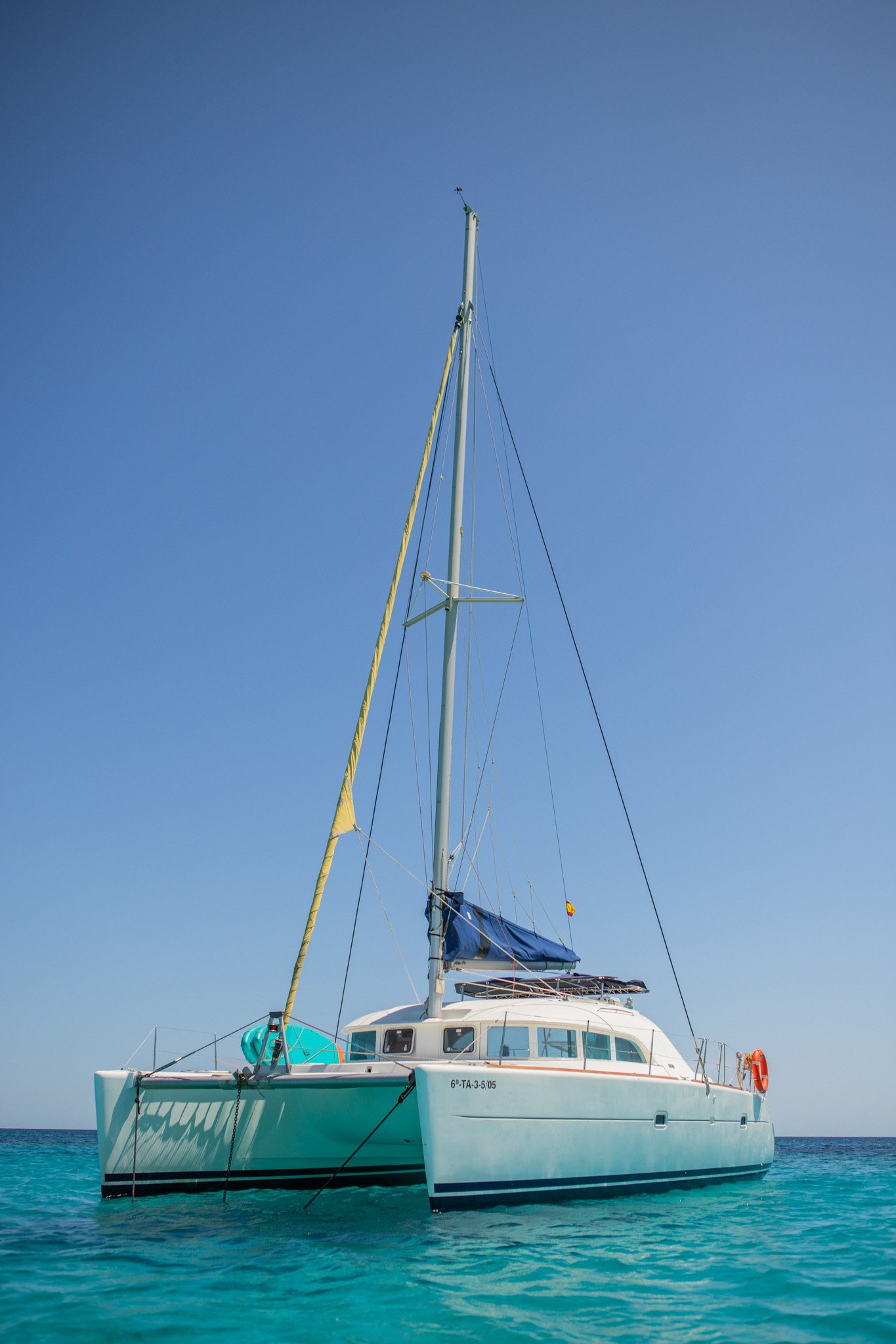 A white yacht with a blue sail cover floating in clear turquoise water, with a tall mast and a clear blue sky in the background.