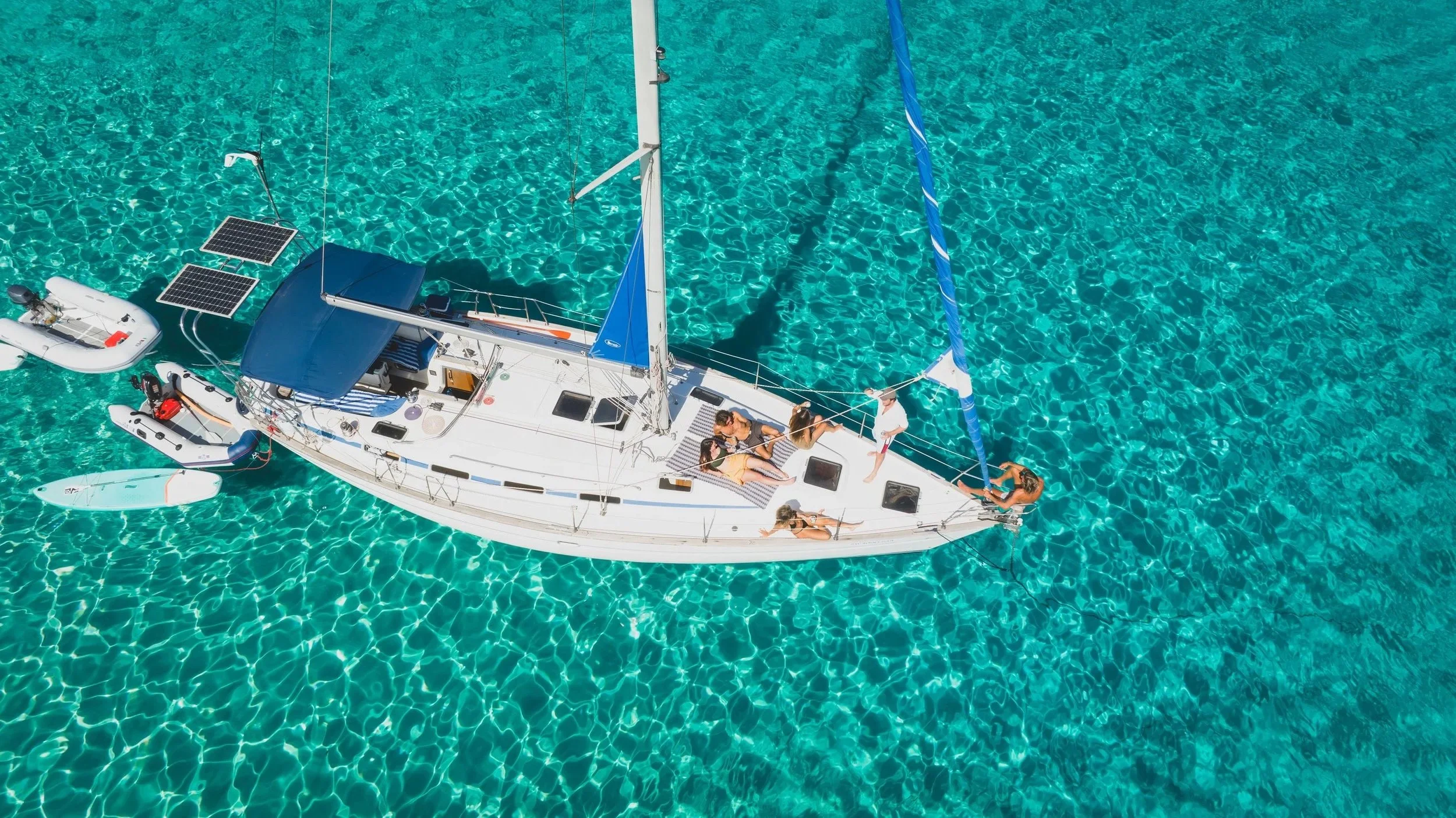 An aerial view of a white sailboat with a blue sail in clear turquoise water. There are five people on the boat, some sitting and others standing, enjoying the sunny day. The boat has solar panels and a small inflatable boat equipped with motors attached to it.