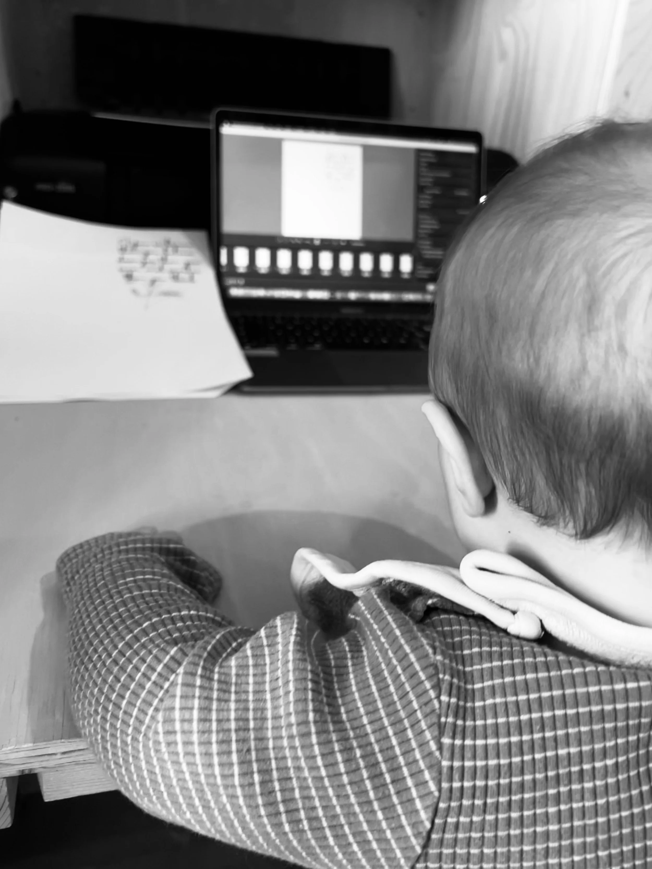 A baby sitting in front of a laptop, with a speaker in the background, and a piece of paper and a box near the laptop.