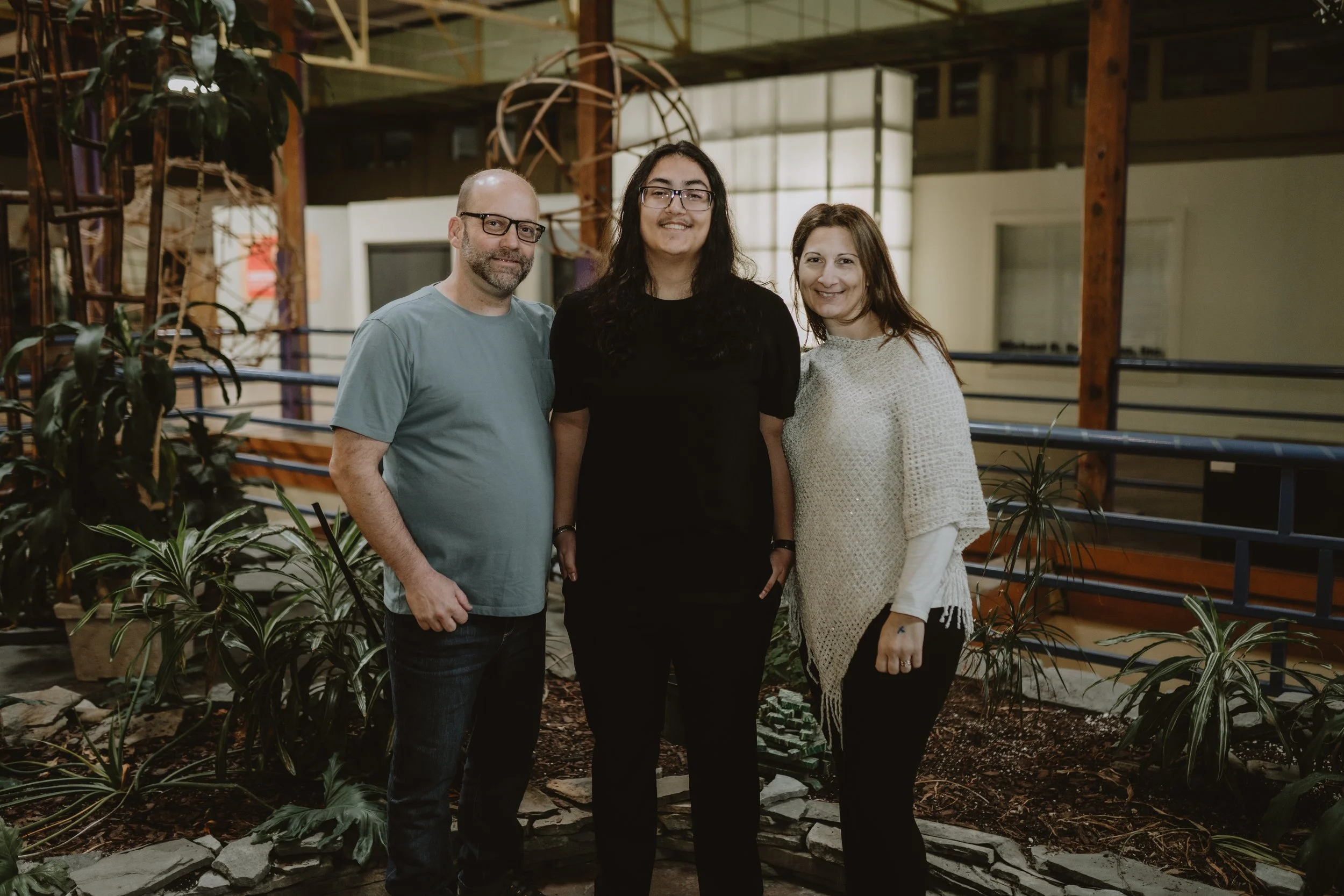Three people standing inside a greenhouse surrounded by plants, with wooden and metal structures in the background.