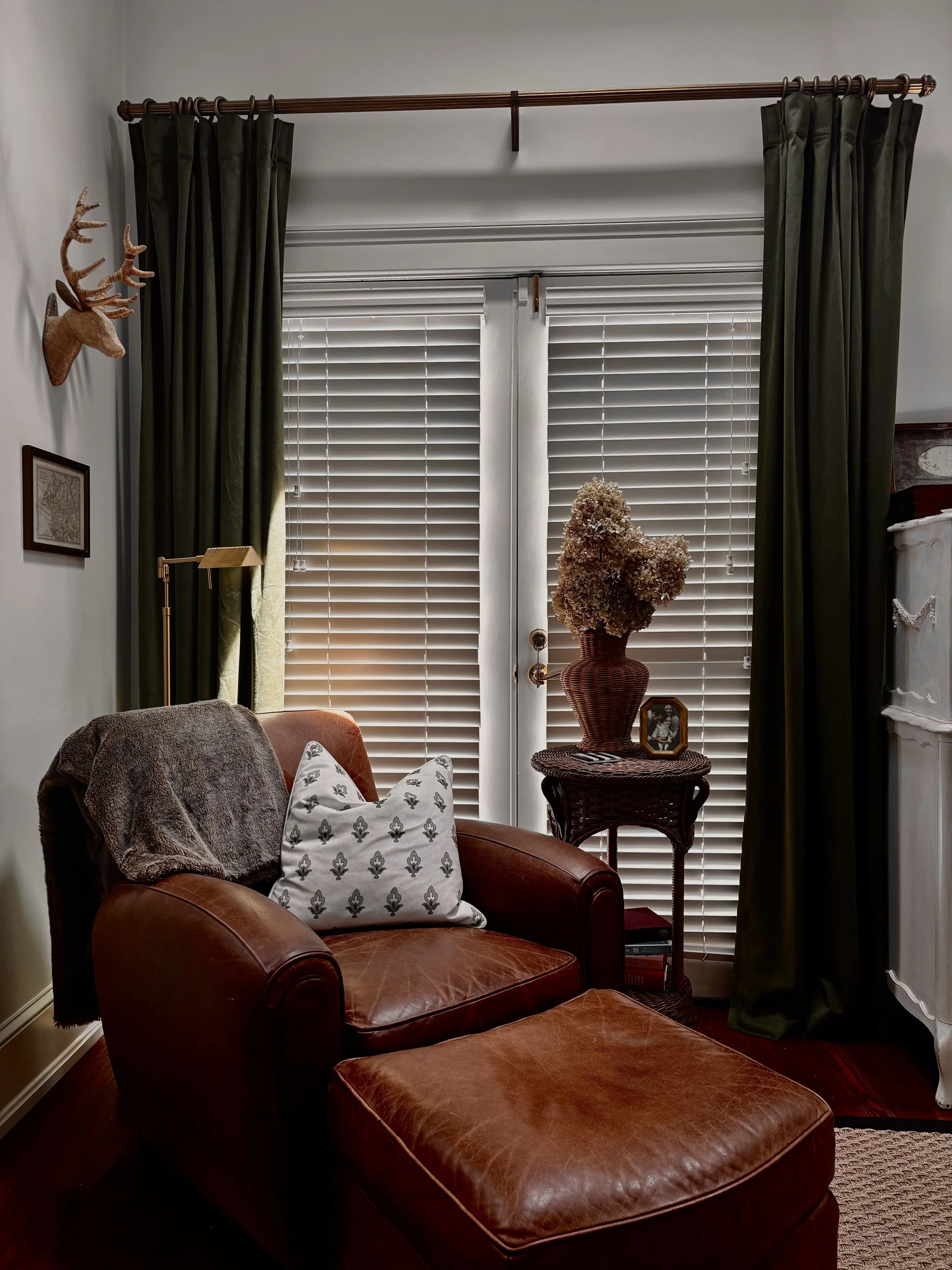 Living room with a leather armchair and footrest, a side table with a framed photograph and dried flowers, green curtains, blinds, and decorative wall mounted reindeer antlers.