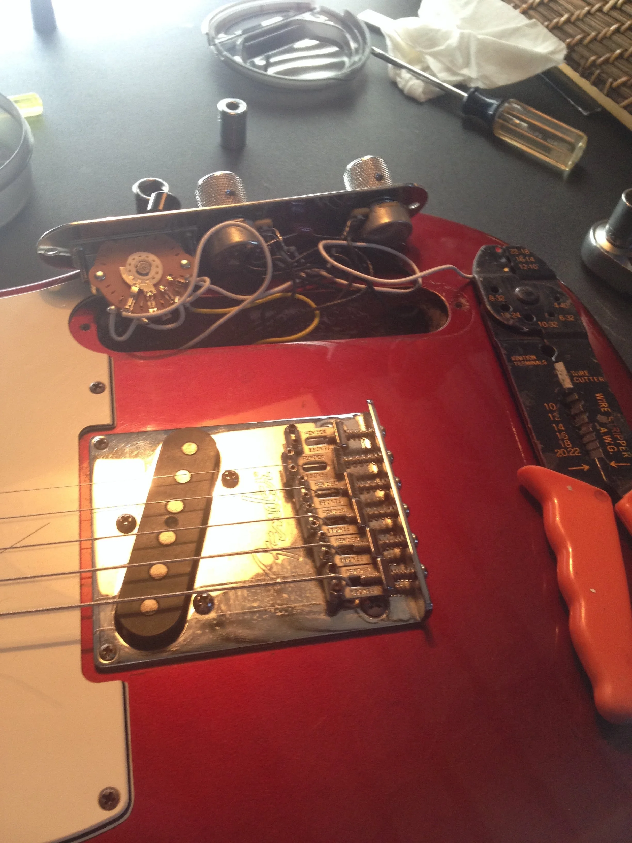 Close-up of an electric guitar with its back cover removed, exposing internal wiring and electronic components, on a red surface with various tools and objects around.