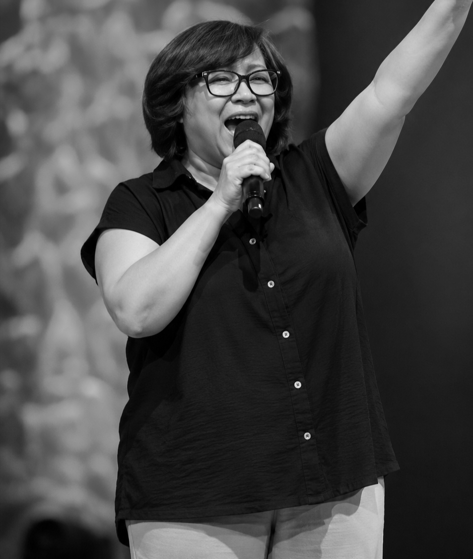 A woman with glasses, wearing a black shirt, smiling and holding a microphone, with her right arm raised, on stage.