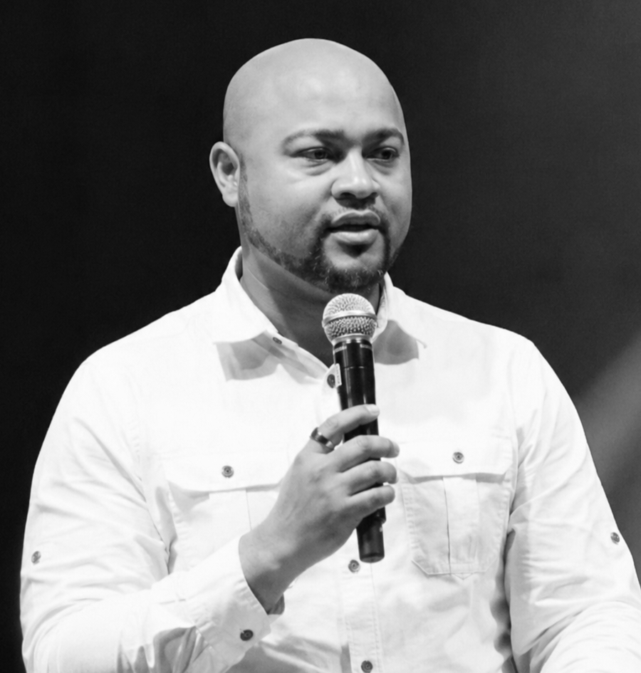 A man with a bald head and beard, wearing a white button-up shirt, holding a microphone and speaking on stage.