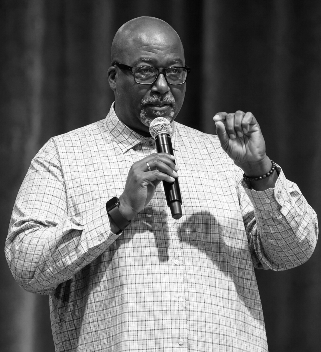 A man with glasses and a beard, wearing a checkered shirt, speaks into a microphone on stage.