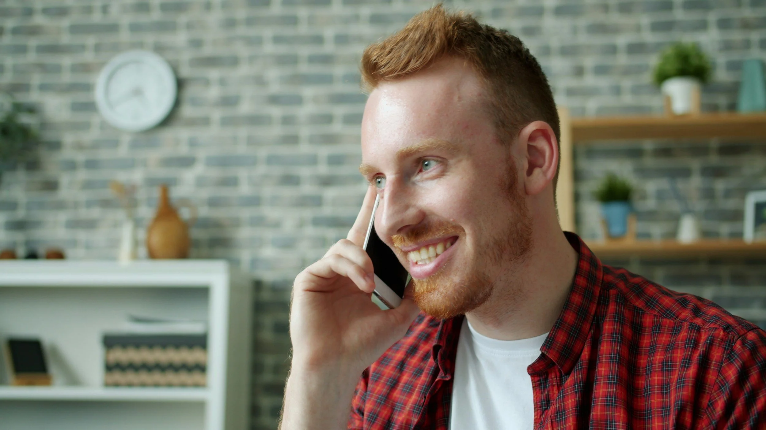 A young man with red hair and a beard, wearing a red plaid shirt, smiling while talking on a cell phone in a modern indoor setting.