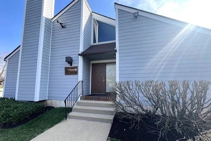 Front entrance of a modern light blue office building with stairs leading up to a wooden door, a black mailbox, and a small window near the door. Bushes and a sidewalk are in front, with sunlight shining from the top right corner.