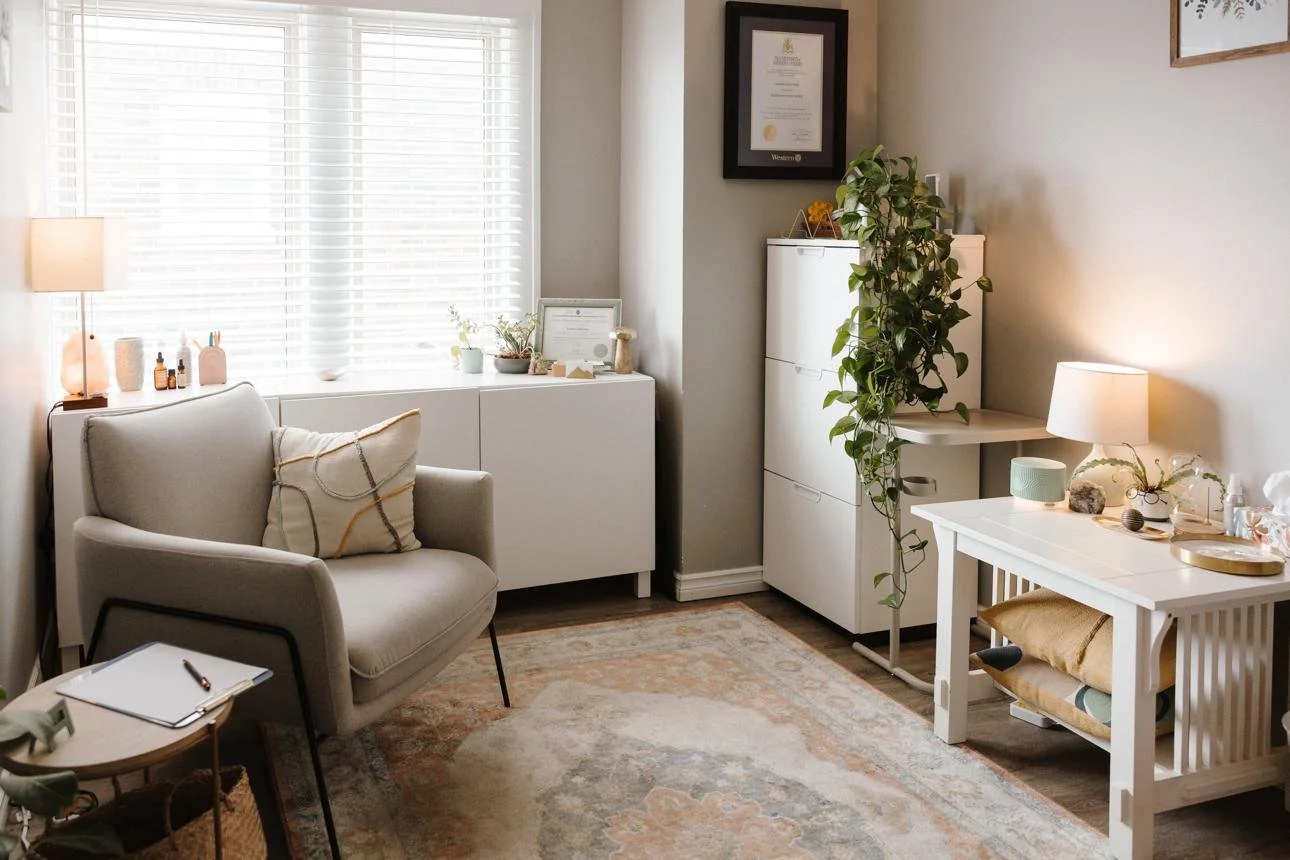 A cozy well-lit therapy office with a window, beige armchair, white cabinet, framed certificate, green plant, white side table with lamp and decor, and assorted decorative items.
