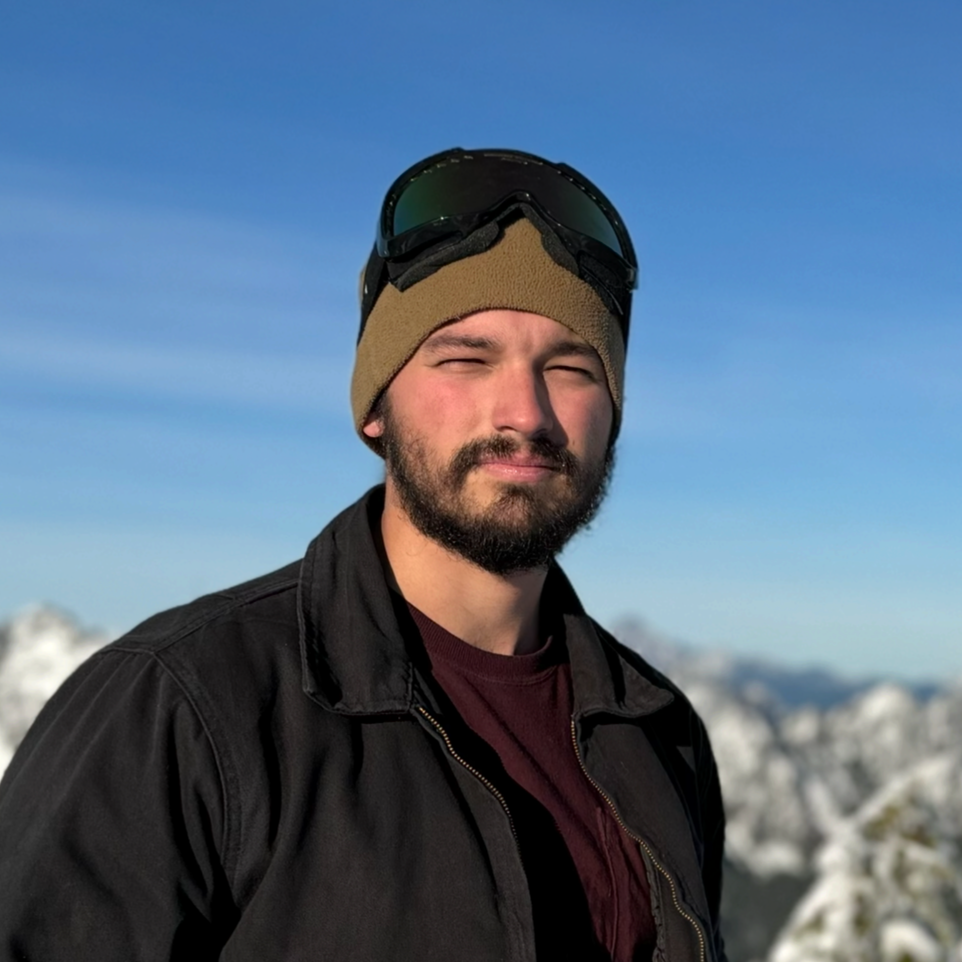 A man outdoors in a snowy mountain landscape, wearing a black jacket, a brown beanie, and ski goggles on his helmet, squinting slightly against the sunlight.