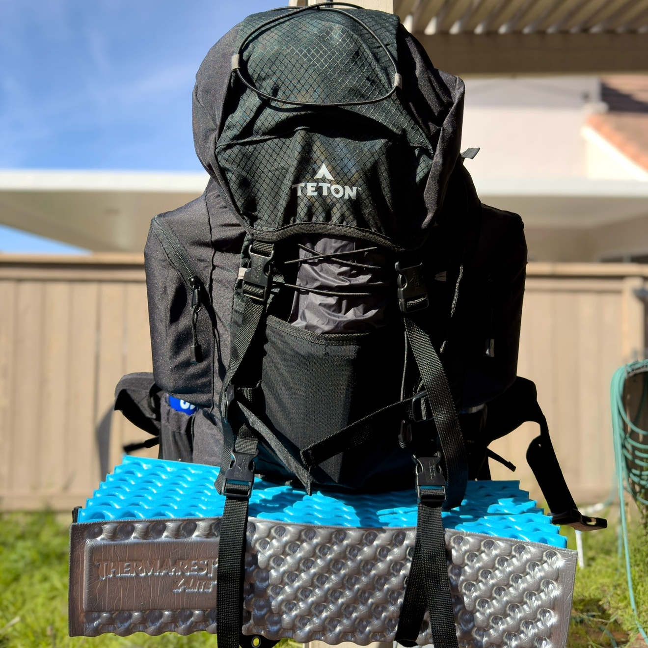 A black Teton backpack placed on a blue and silver foam pad outdoors, with a wooden fence and house in the background.