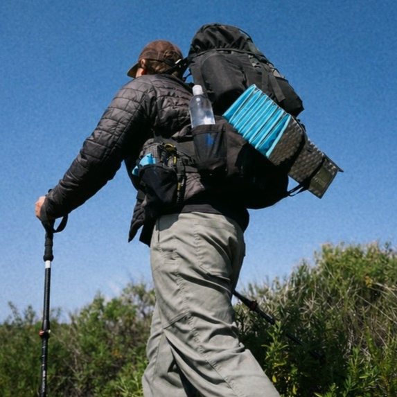 Person hiking with a large backpack and trekking poles on a sunny day.