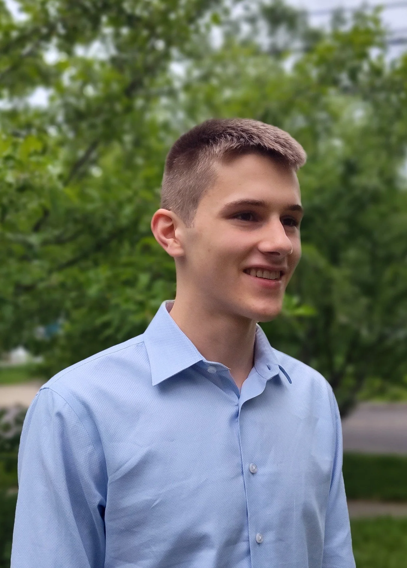 A young man with short brown hair and light skin, smiling, wearing a light blue dress shirt, standing outdoors with green trees in the background.