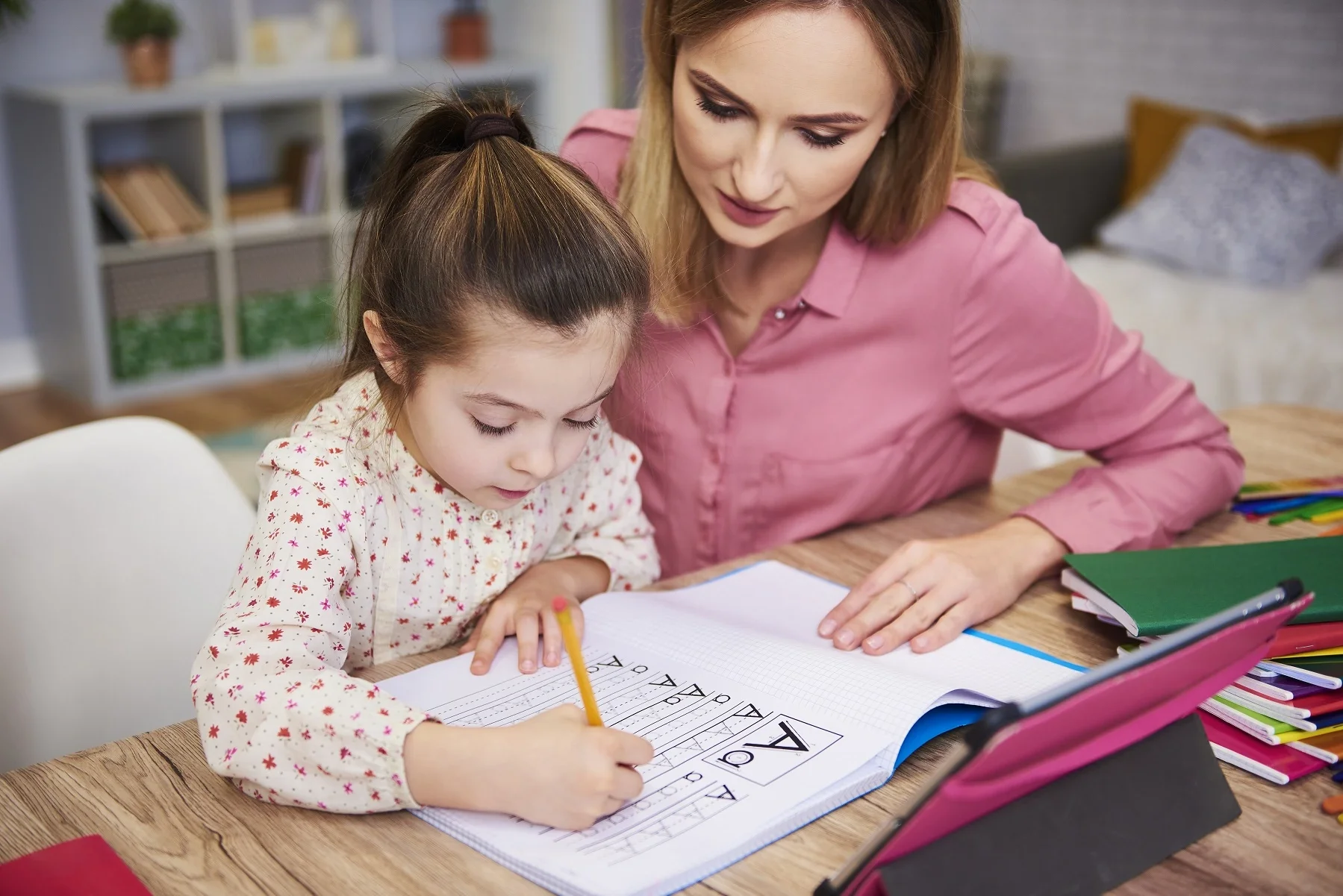 A woman and a young girl doing homework together at a wooden table. The girl is writing in a notebook, and the woman is guiding her. The table has colored pencils and notebooks on it.