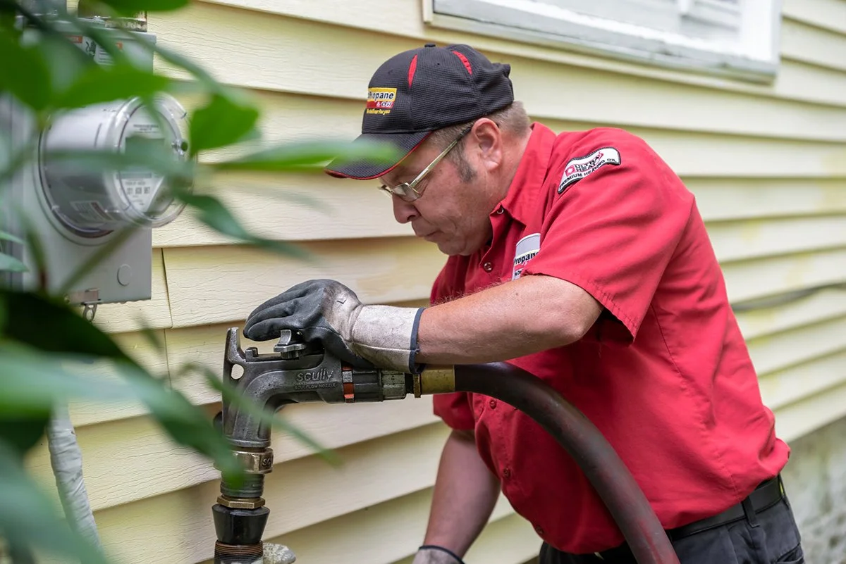 A man in a red uniform and black cap is working on outdoor plumbing with a wrench, next to a yellow house and a utility meter.