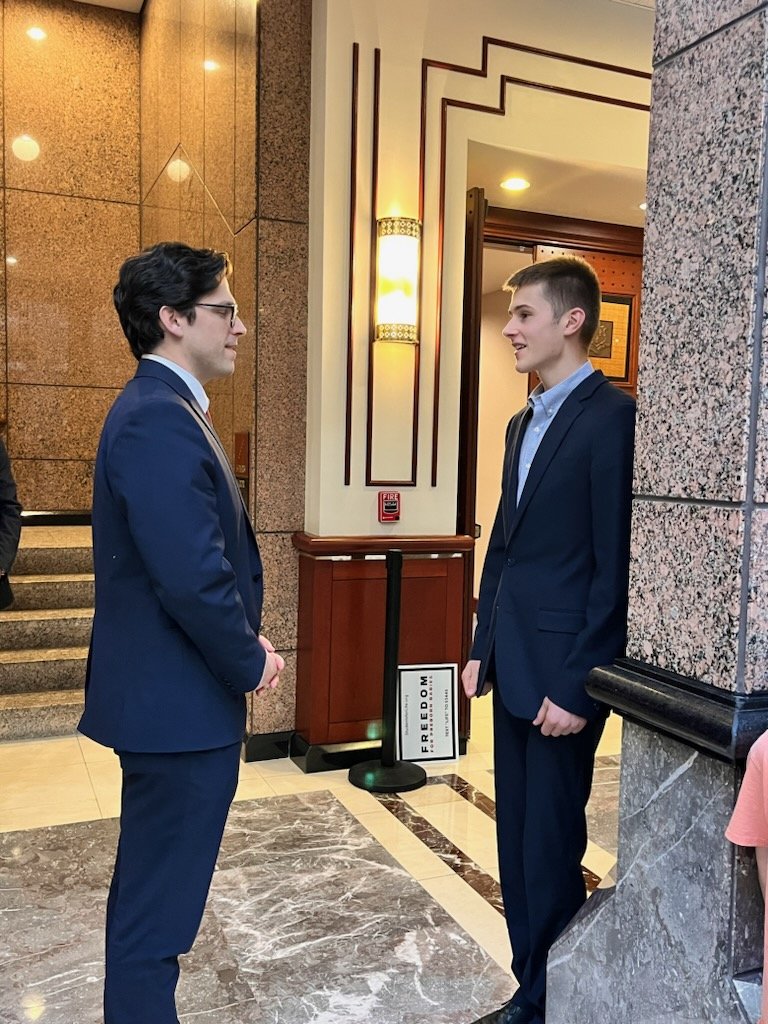 Two young men in suits standing face to face, chatting in an indoor setting with marble flooring and decorative wall panels.