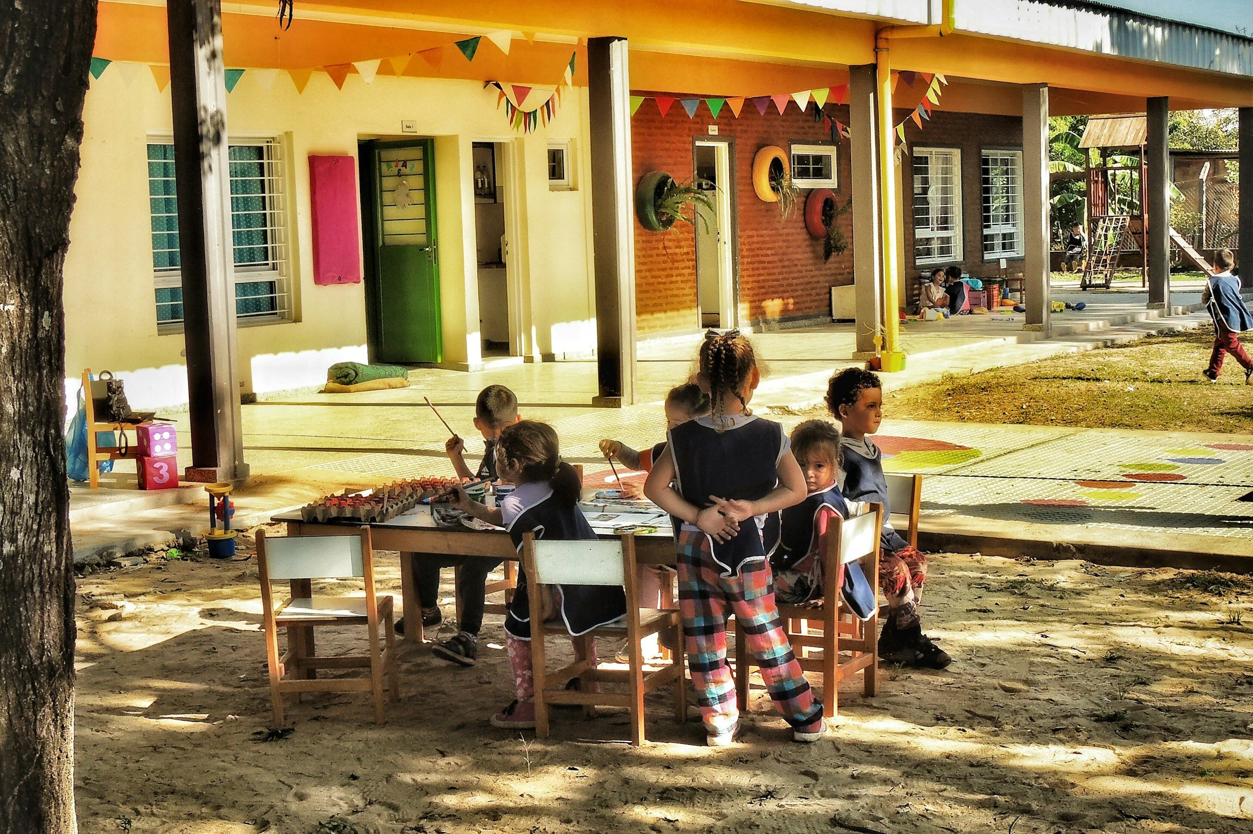 Children playing and sitting outside a colorful building, possibly a kindergarten or preschool, decorated with bunting flags, with playground equipment visible.