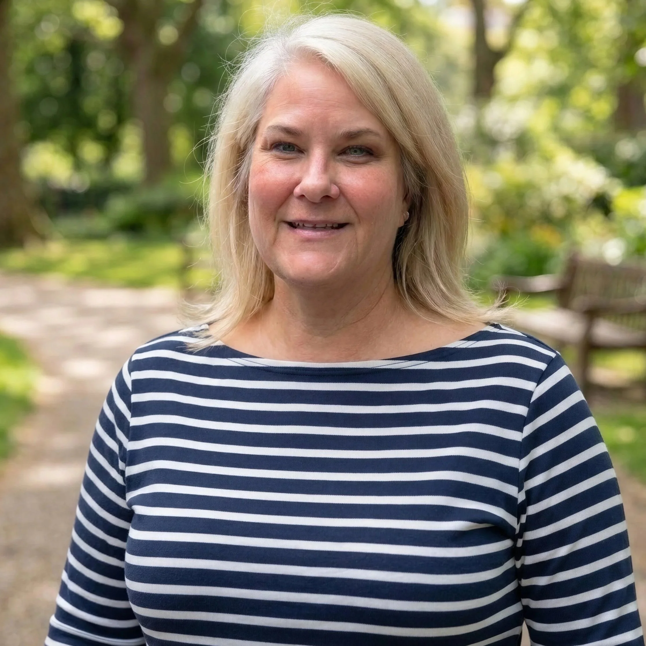 Close-up of a smiling middle-aged woman with blonde hair, wearing a navy and white striped shirt, standing outdoors with green trees and a pathway in the background.