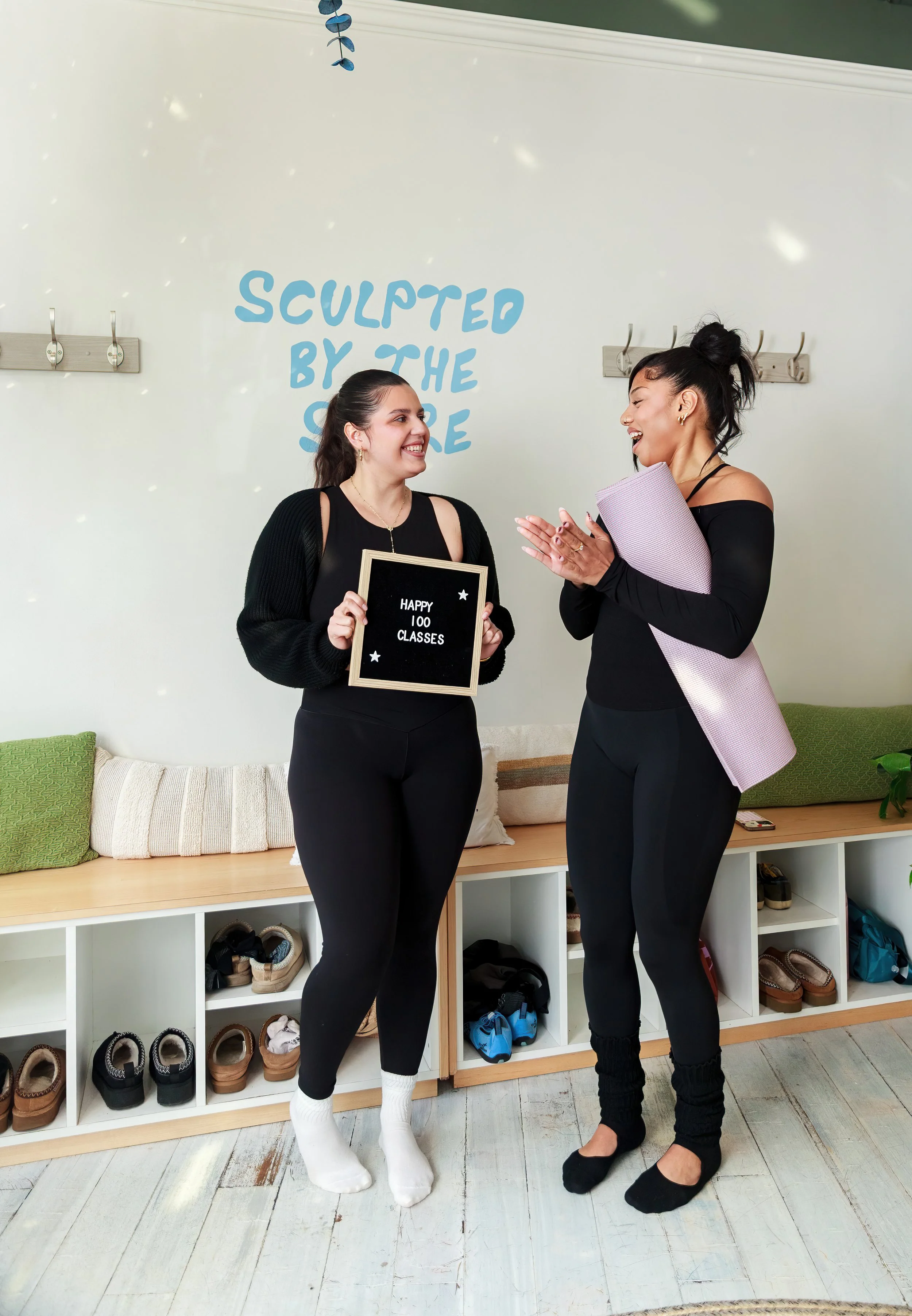 Two women smiling and chatting indoors. One woman is holding a sign that says 'Happy 100 Classes,' and the other woman is holding a pink yoga mat. They are standing in front of a white wall with the phrase 'Sculpted by the Scope' written on it. There is a white shoe rack with multiple pairs of shoes beneath them.