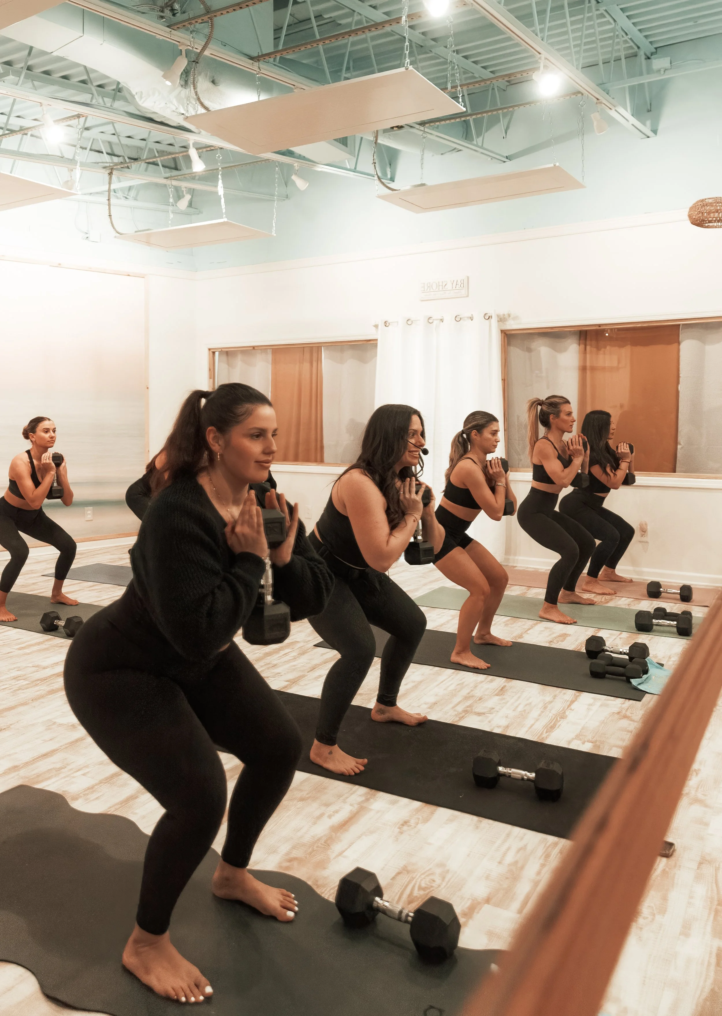 Group of women doing squats with dumbbells in a yoga class, standing on black mats in a room with wooden floors and mirror windows.