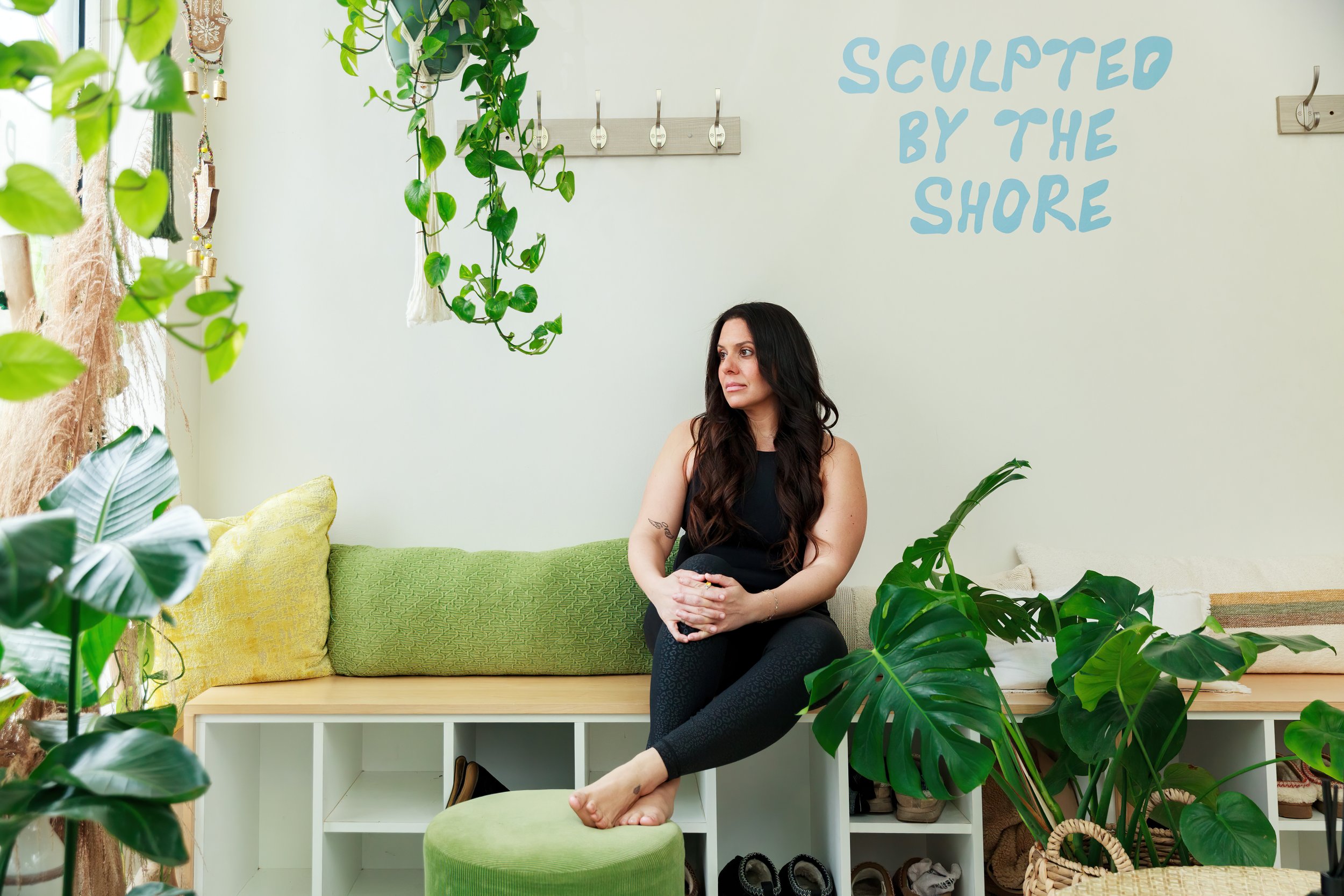 A woman sitting on a green bench in a room filled with various green plants. She is wearing a black sleeveless top and black leggings, with her hands clasped in her lap. Behind her, there are colorful pillows on a green couch and a white wall with blue text that reads 'SCULPTED BY THE SHORE.'