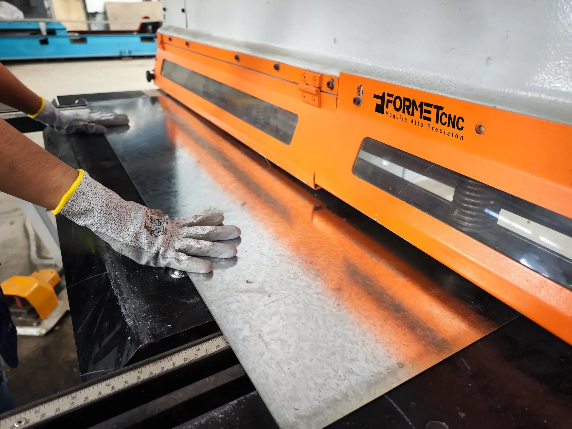 Workers handling a sheet of metal using a precision cutting or shaping machine, with orange and black safety gear in an industrial setting.