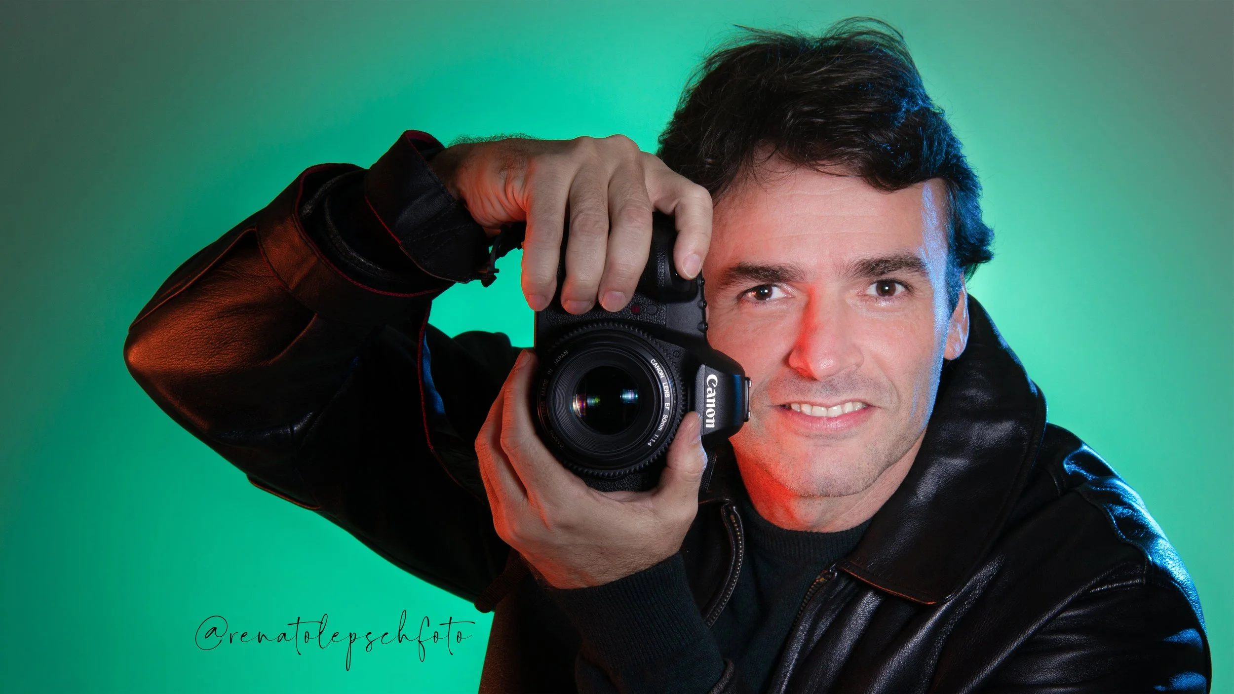 Homem com cabelo escuro, vestindo jaqueta de couro preta, segurando uma câmera Canon apontada para a câmera, sorrindo em fundo verde com iluminação de destaque